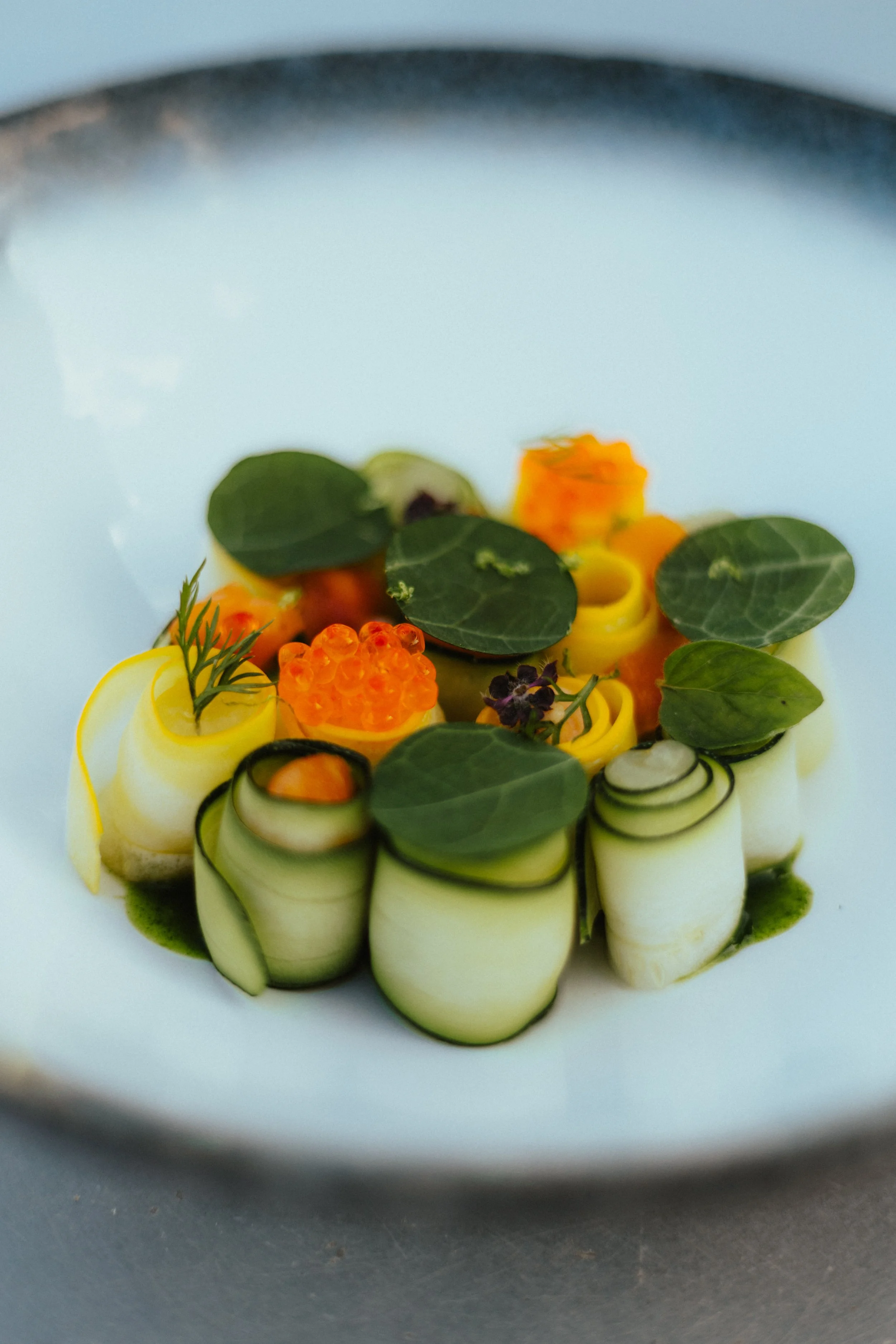 Assiette de tartare de légumes décorée avec des feuilles vertes, des petites fleurs, et de la gelée orange, présentée de manière artistique.