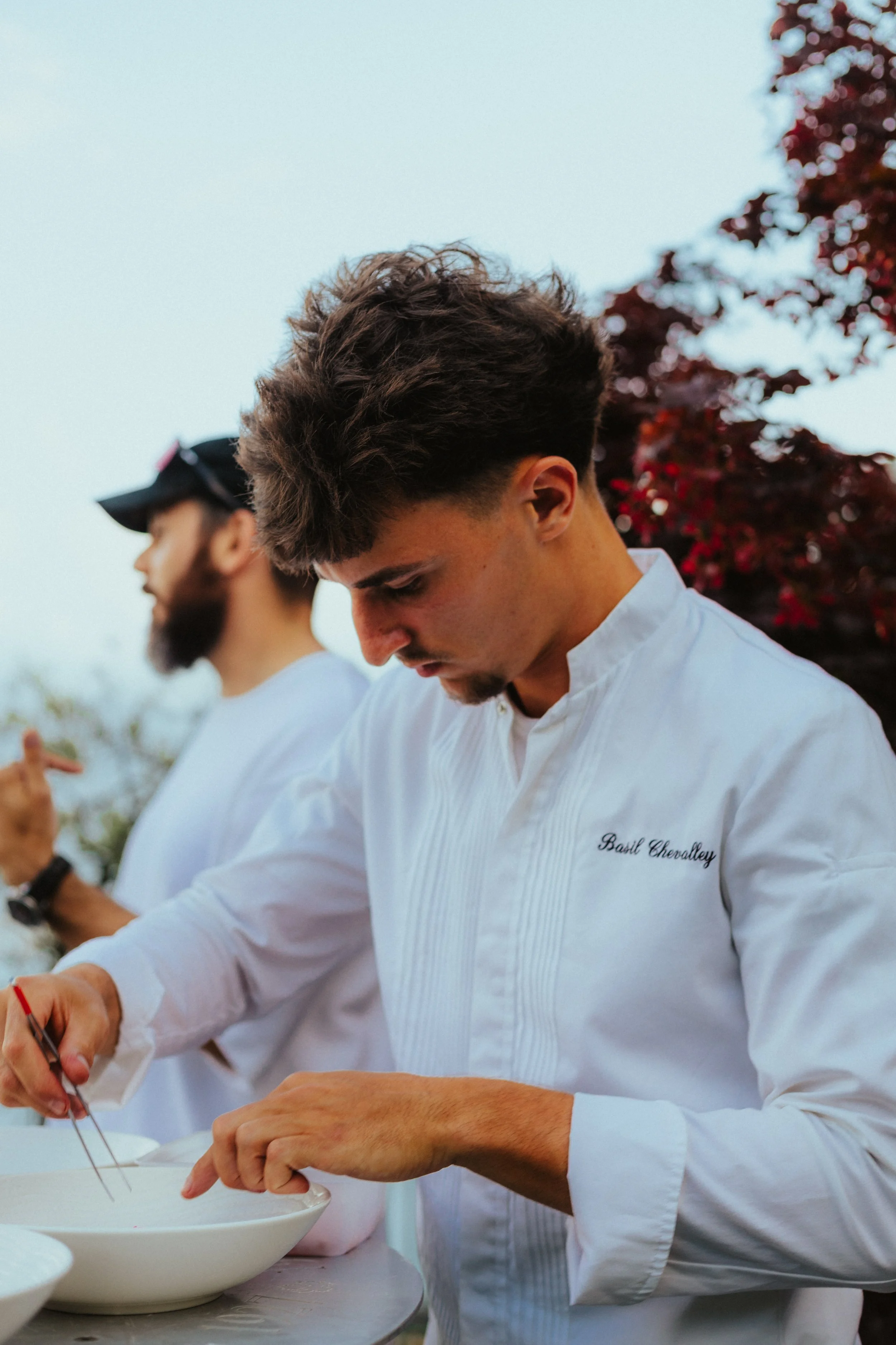 Deux hommes en blanc, dont un porte un nom brodé 'Basil Chevalley', préparent ou dressent des plats dans un espace extérieur, avec un arbre aux feuilles rouges en arrière-plan.