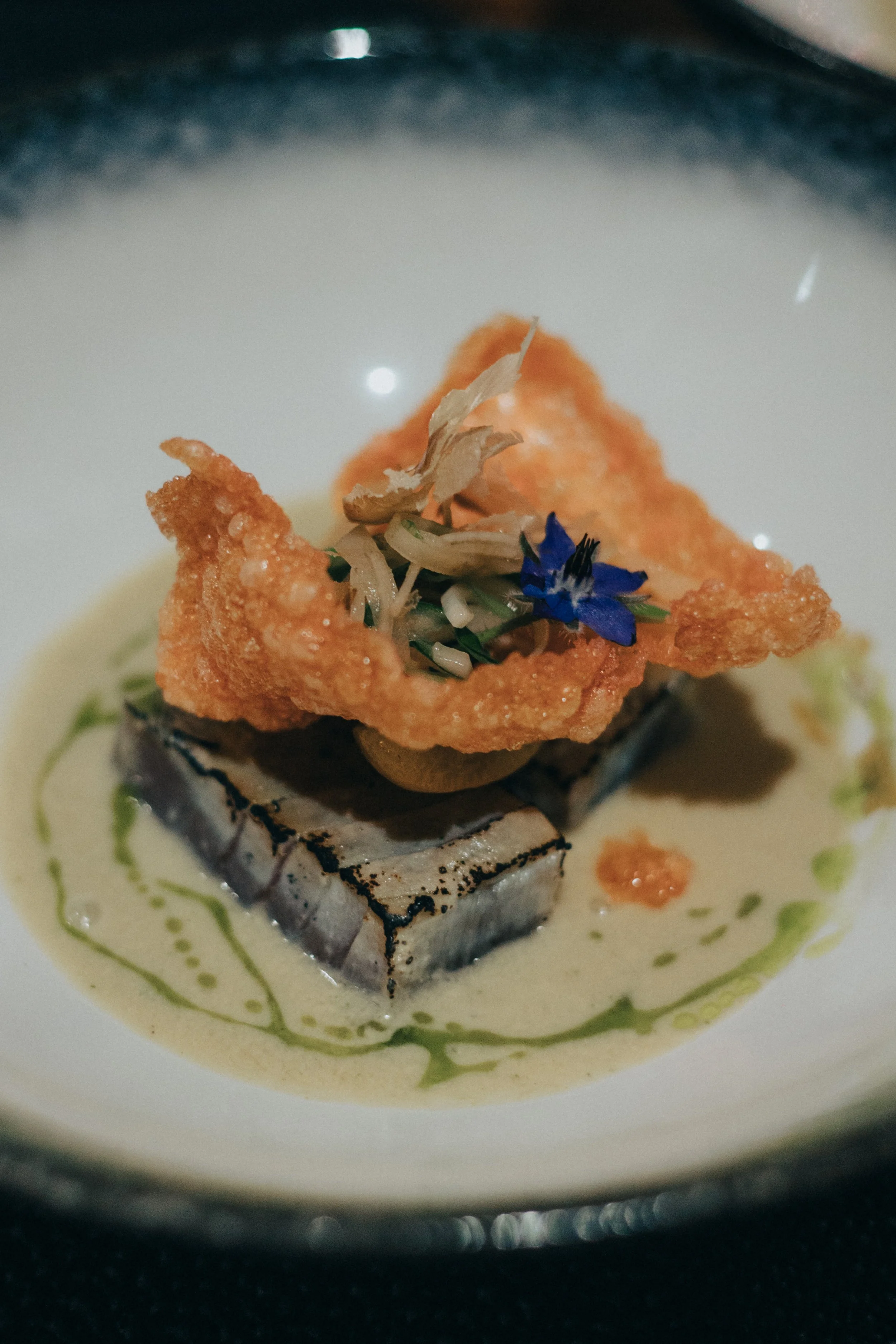 Plat gastronomique avec un morceau de poisson grillé, une friture croustillante, une garniture d'herbes et fleurs comestibles, dans une sauce crémeuse.