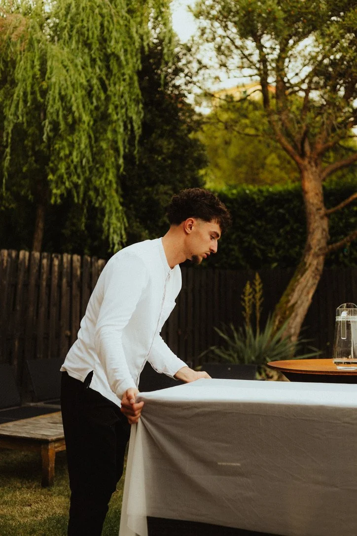 Un jeune homme en chemise blanche ajuste une nappe sur une table à l'extérieur dans un jardin, avec des arbres en arrière-plan.