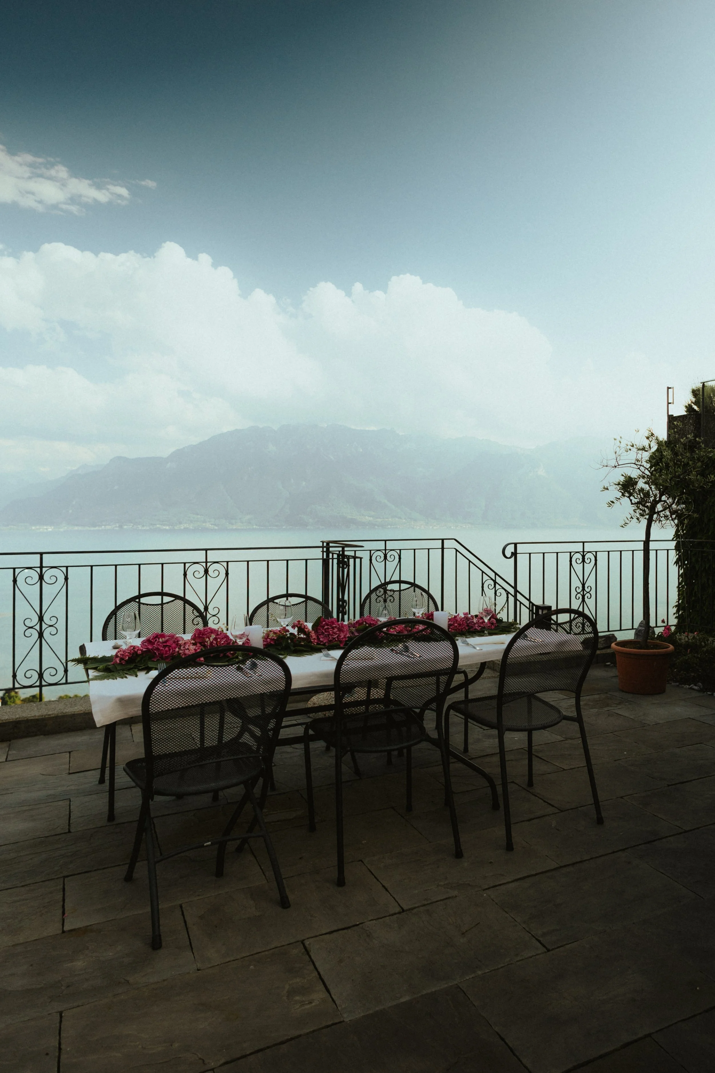 Table dressée avec des chaises en terrasse avec vue sur un lac et des montagnes, décorée de fleurs roses.