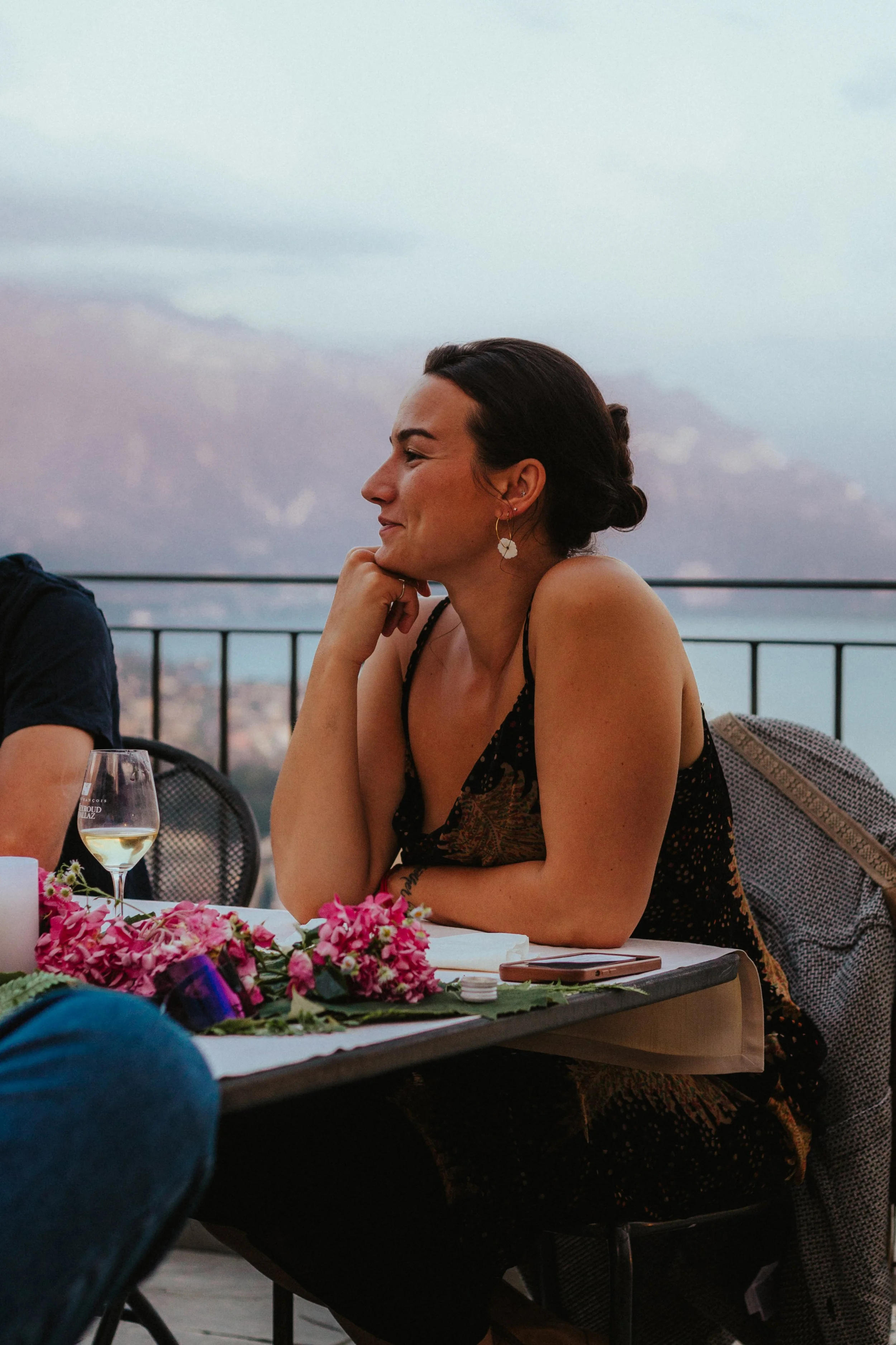 Femme assise à une table lors d'un dîner en plein air, avec un verre de vin blanc et des fleurs roses sur la table, regardant vers la gauche, avec une vue sur la mer en arrière-plan.
