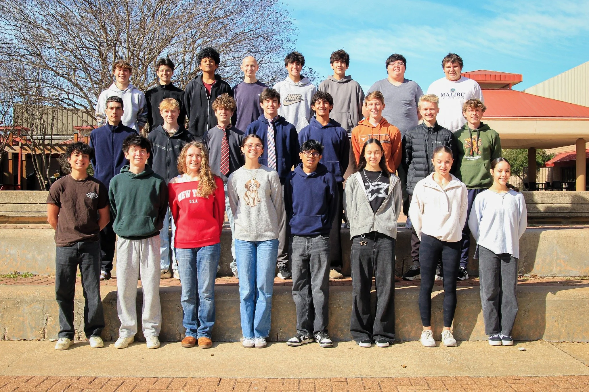Group of students standing outdoors on steps, posing for a class photo on a sunny day with a clear blue sky and trees in the background.
