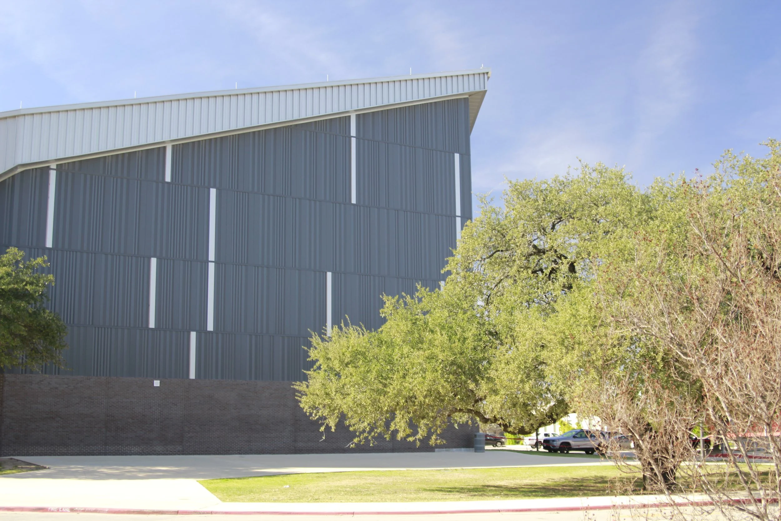 Side view of a modern building with vertical metal siding, large trees with green foliage in the foreground, parking lot with cars, and a blue sky with some clouds.