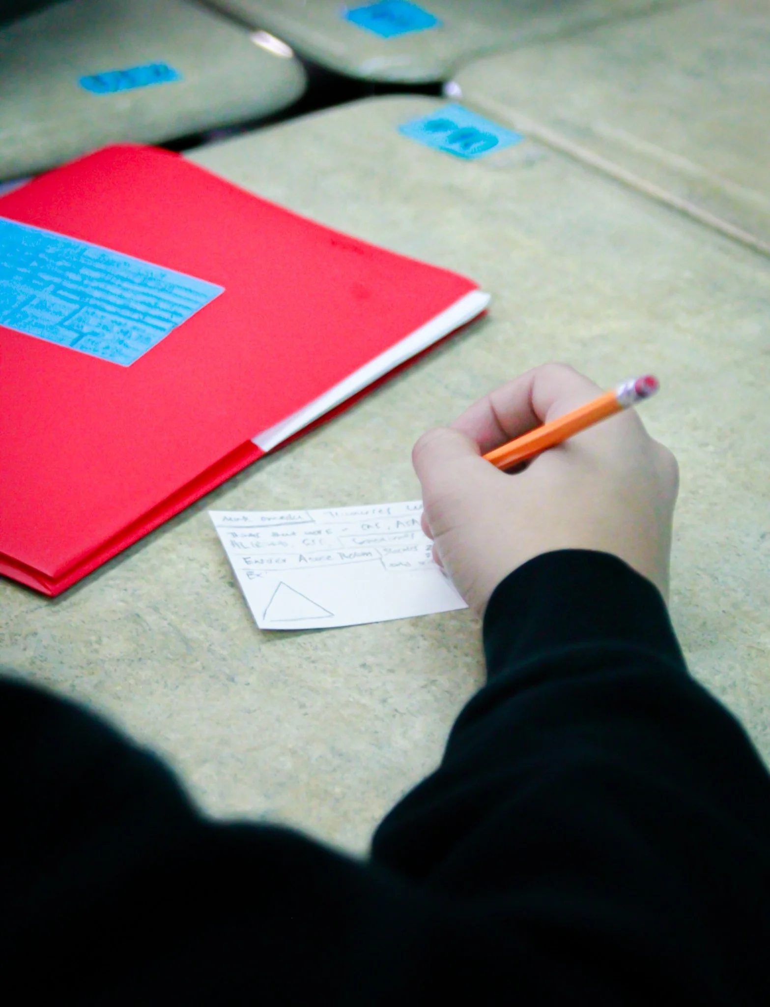 A person wearing a black long-sleeve shirt writing on a small piece of paper with a pencil, with a red notebook and other folders on a beige desk.