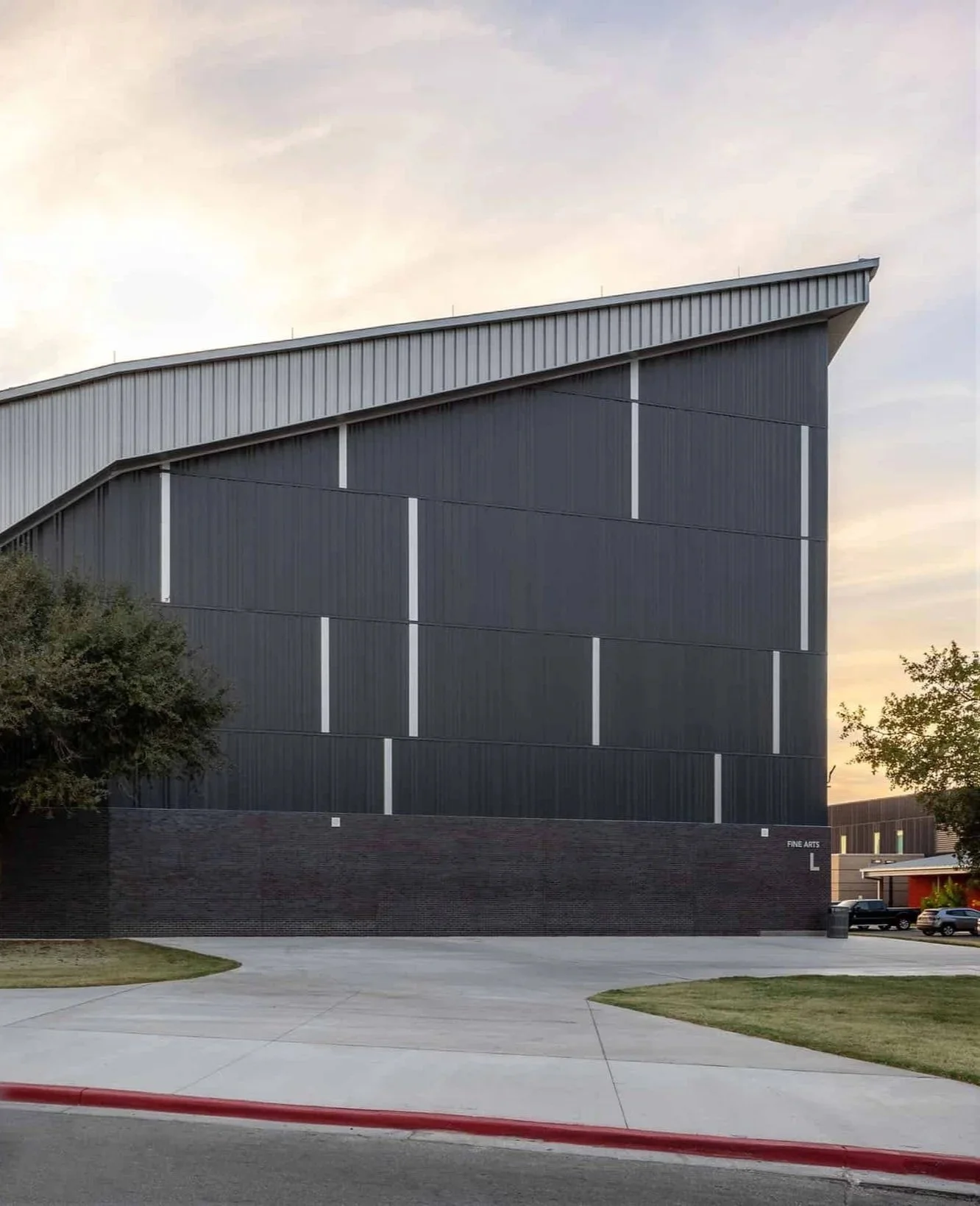 A modern building with dark exterior walls and a sloped roof, with a small lawn, sidewalk, trees, and a parking lot nearby at sunset.
