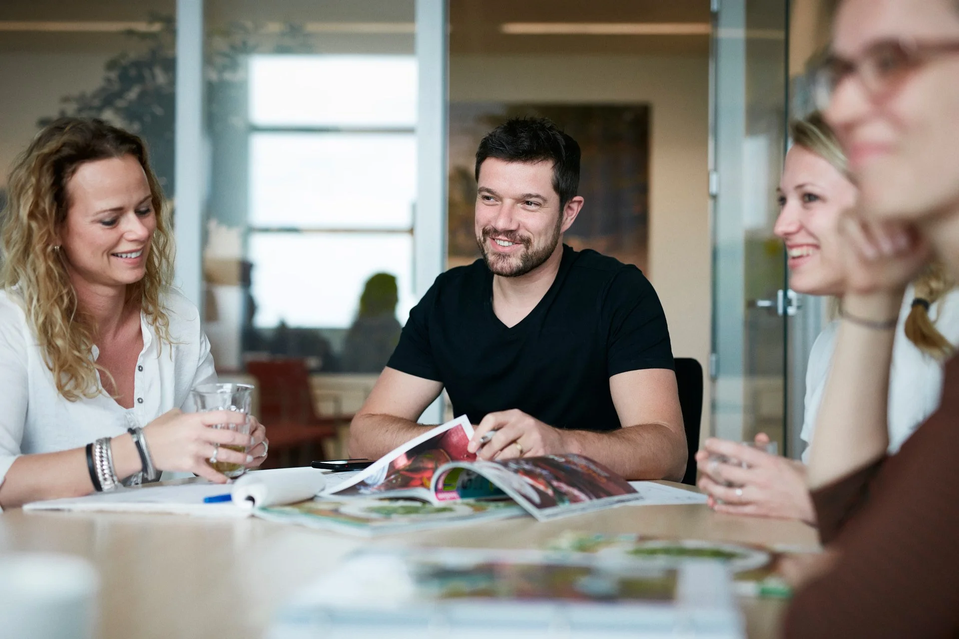Group of four adults sitting at a table, looking at magazines and smiling, in a bright, modern office or meeting room.