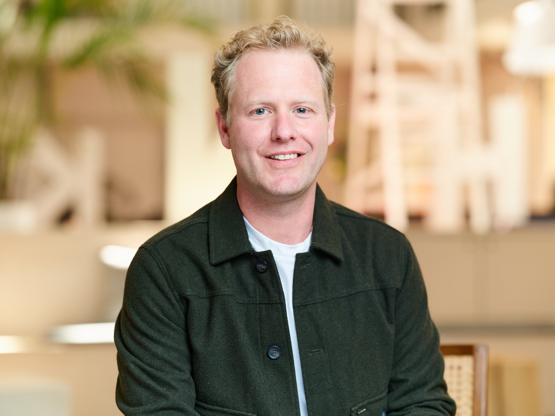 A smiling man with curly blonde hair, wearing a black jacket and white shirt, sitting in a warmly lit room with blurred background.