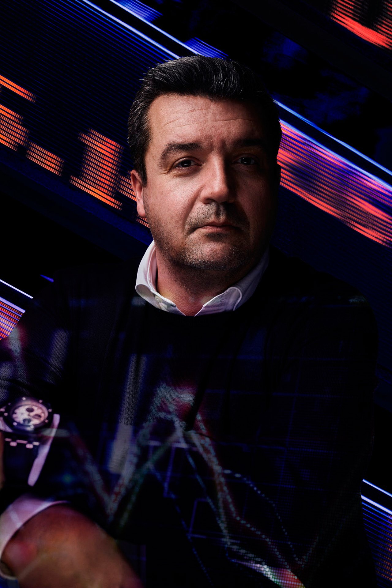 A man with dark hair and a serious expression, wearing a dark jacket and white shirt, is set against a background featuring colorful digital or light streaks in blue, red, and purple hues.