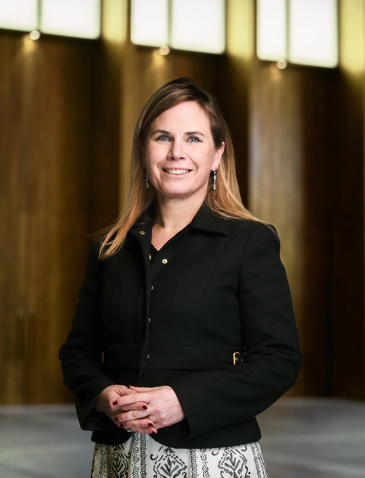 A woman wearing a black blazer and patterned skirt standing inside a building with large windows.