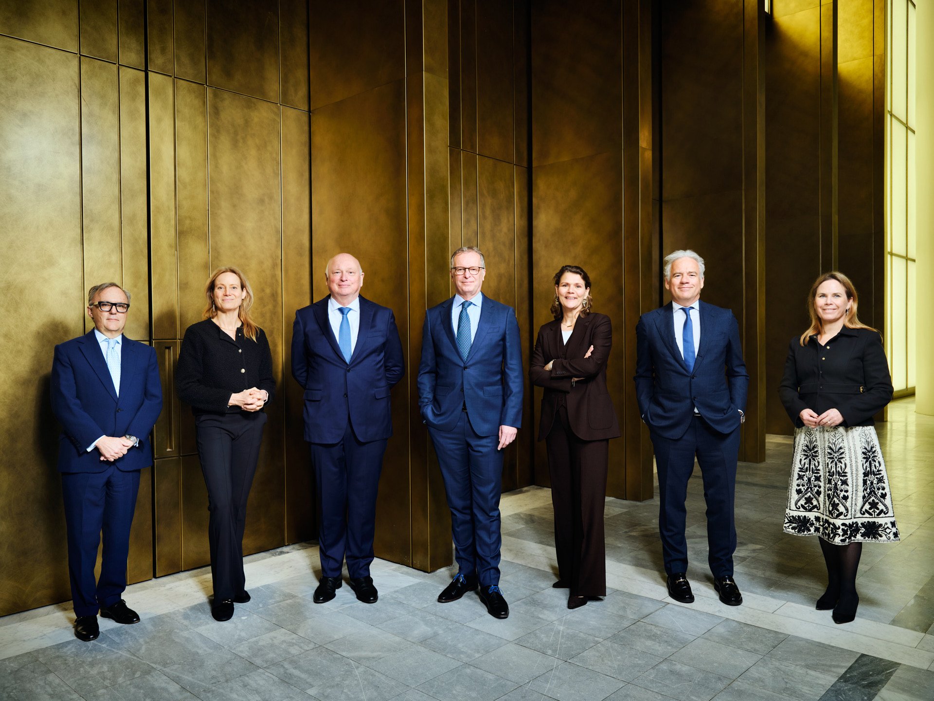 A group of seven professionals, four men and three women, standing in a line in front of a large golden wall in a modern interior.