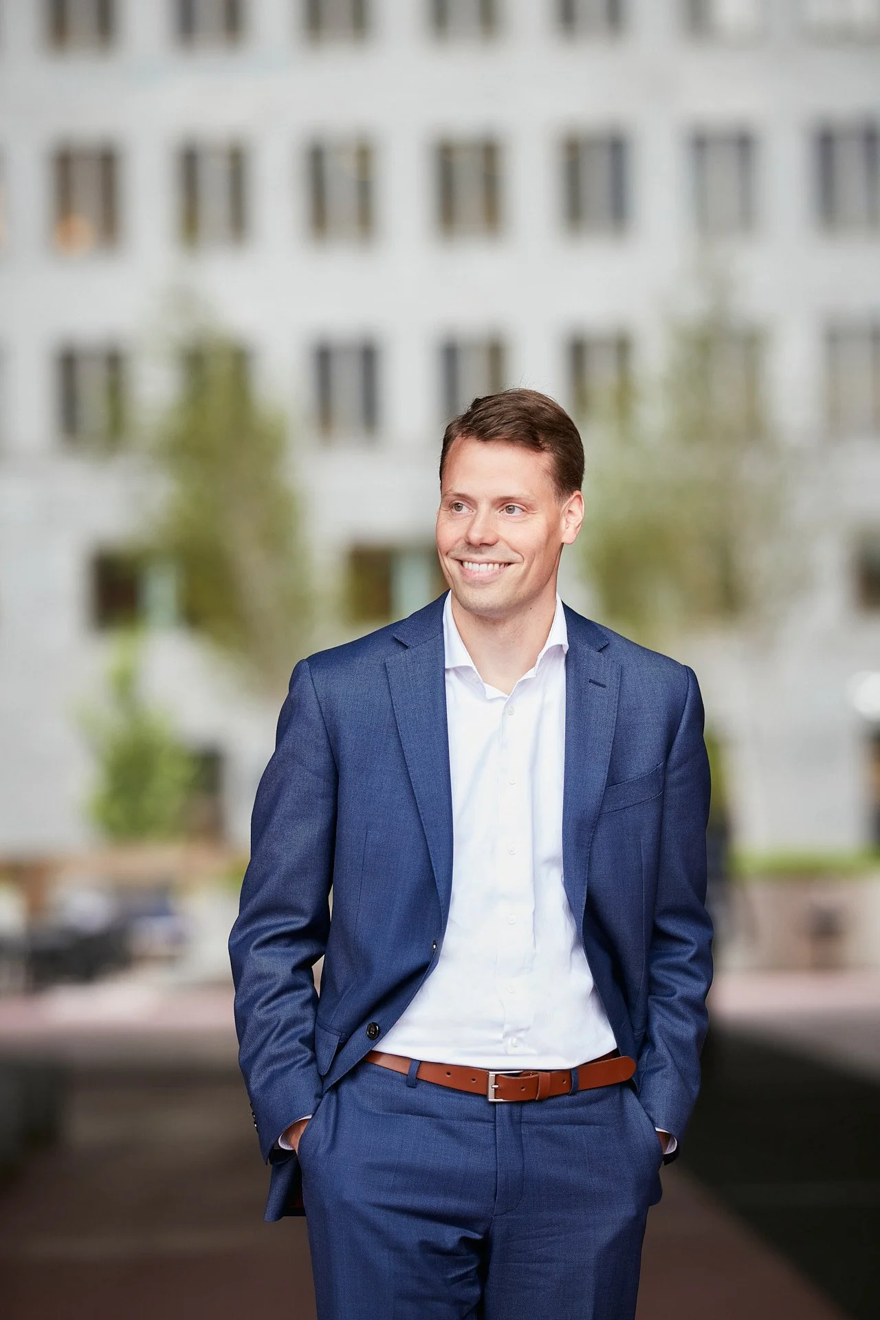 A man in a blue suit with a white shirt and brown belt standing outdoors, smiling, with a blurred background of a modern building with multiple windows and trees.