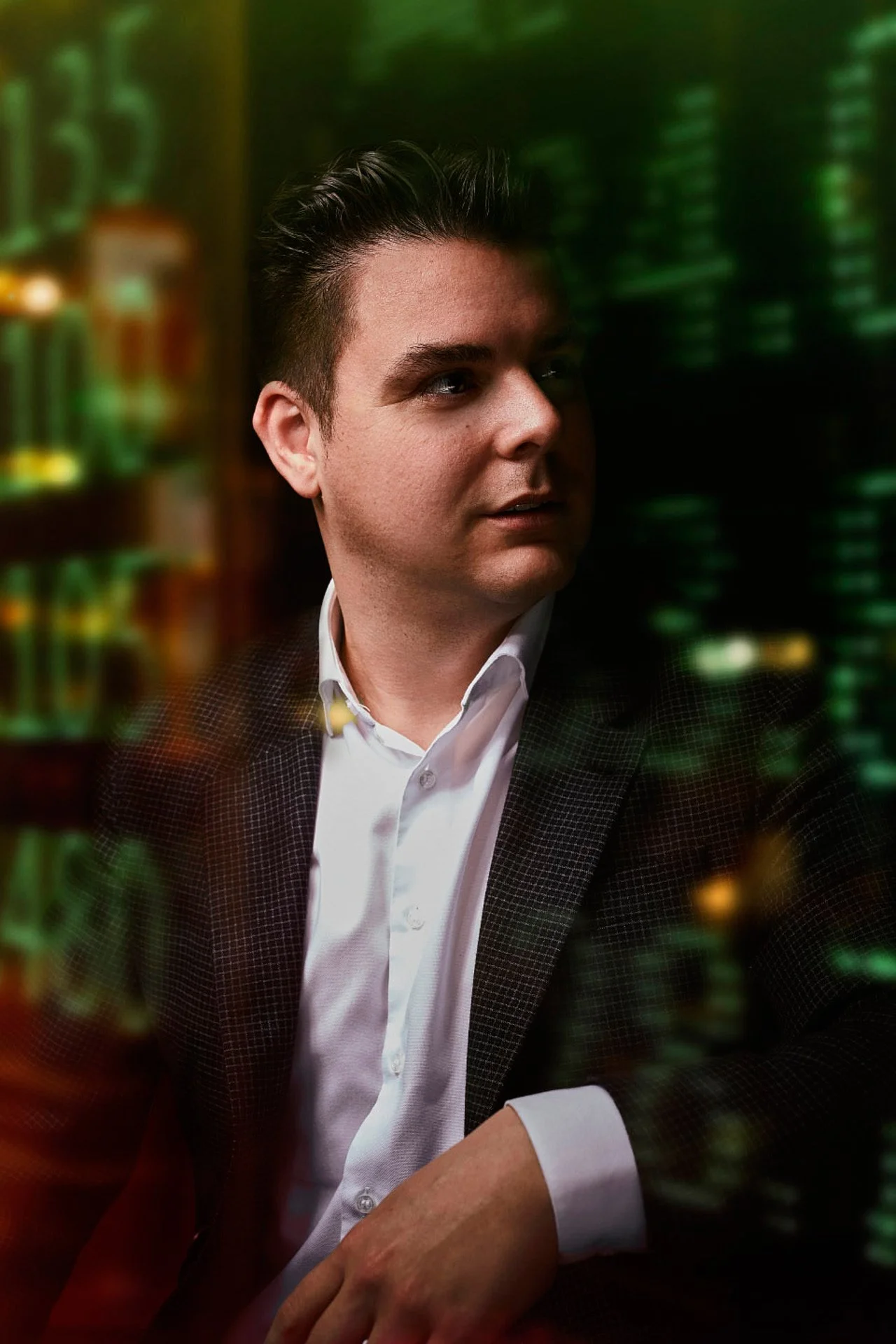 A man dressed in a white shirt and black blazer sitting in front of a blurred data screen with green lights, possibly in a server room or data center.