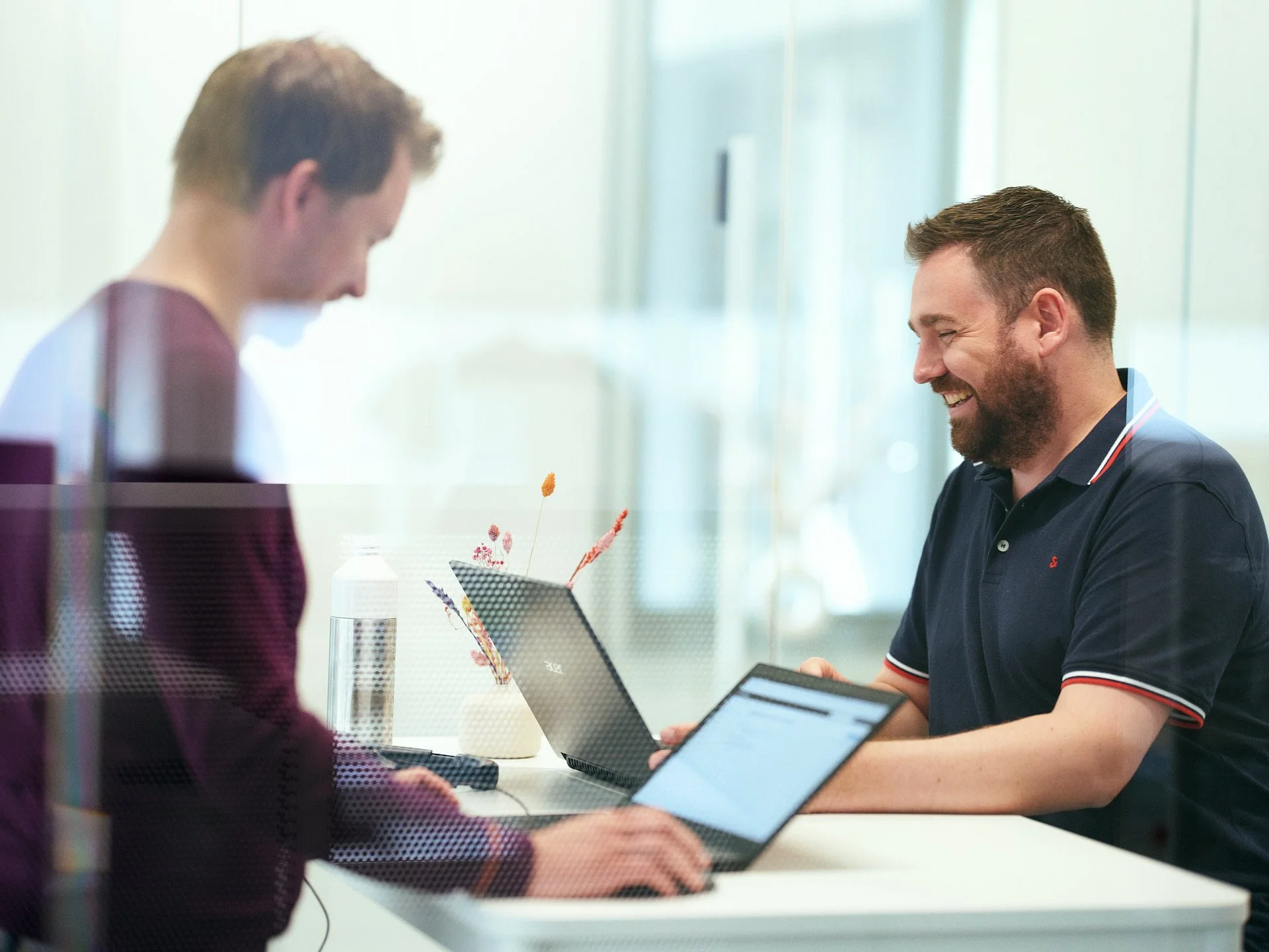 Two men sitting at a white table in a bright office, looking at a laptop and a tablet, smiling and engaging in conversation.