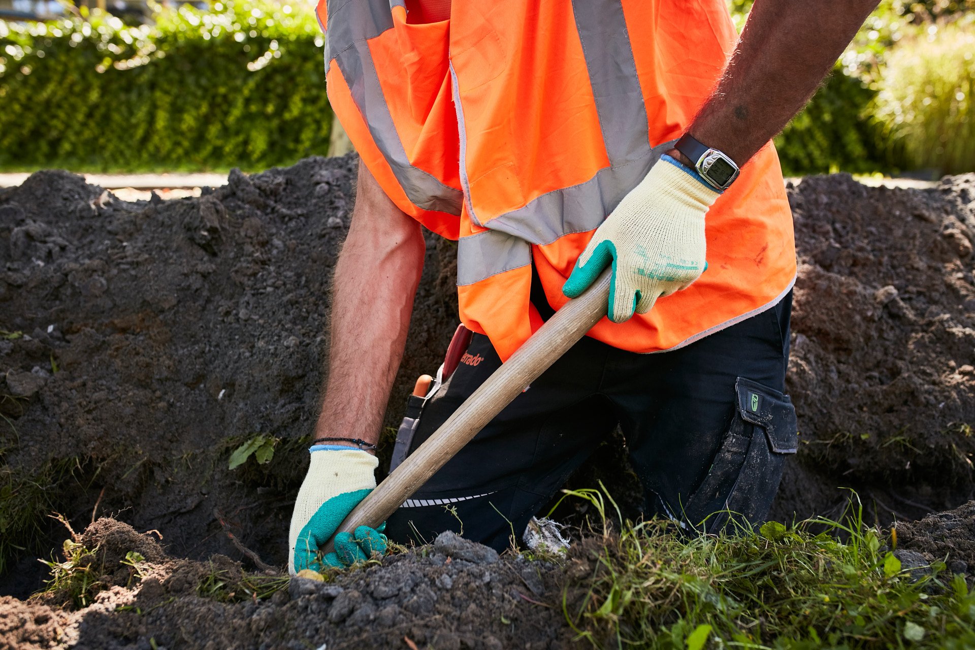 A person wearing orange safety vest, black work pants, and gloves using a shovel to dig in the soil outdoors, with green shrubs in the background.
