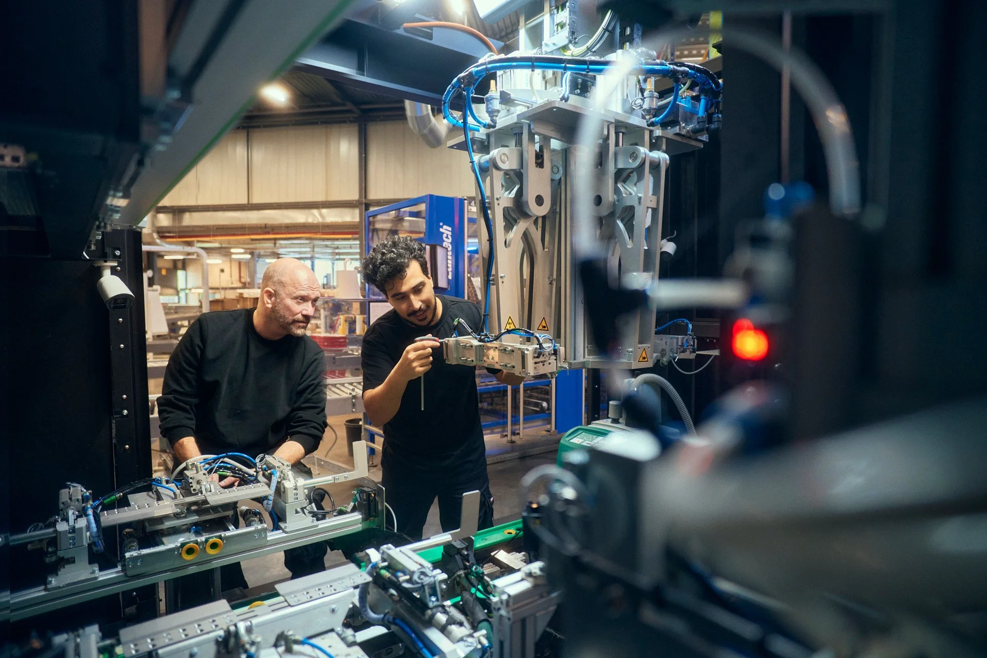 Two men working on industrial machinery in a factory or workshop. One man, with a beard and black shirt, looks on as the other, with curly hair and headset, adjusts a component of the equipment.