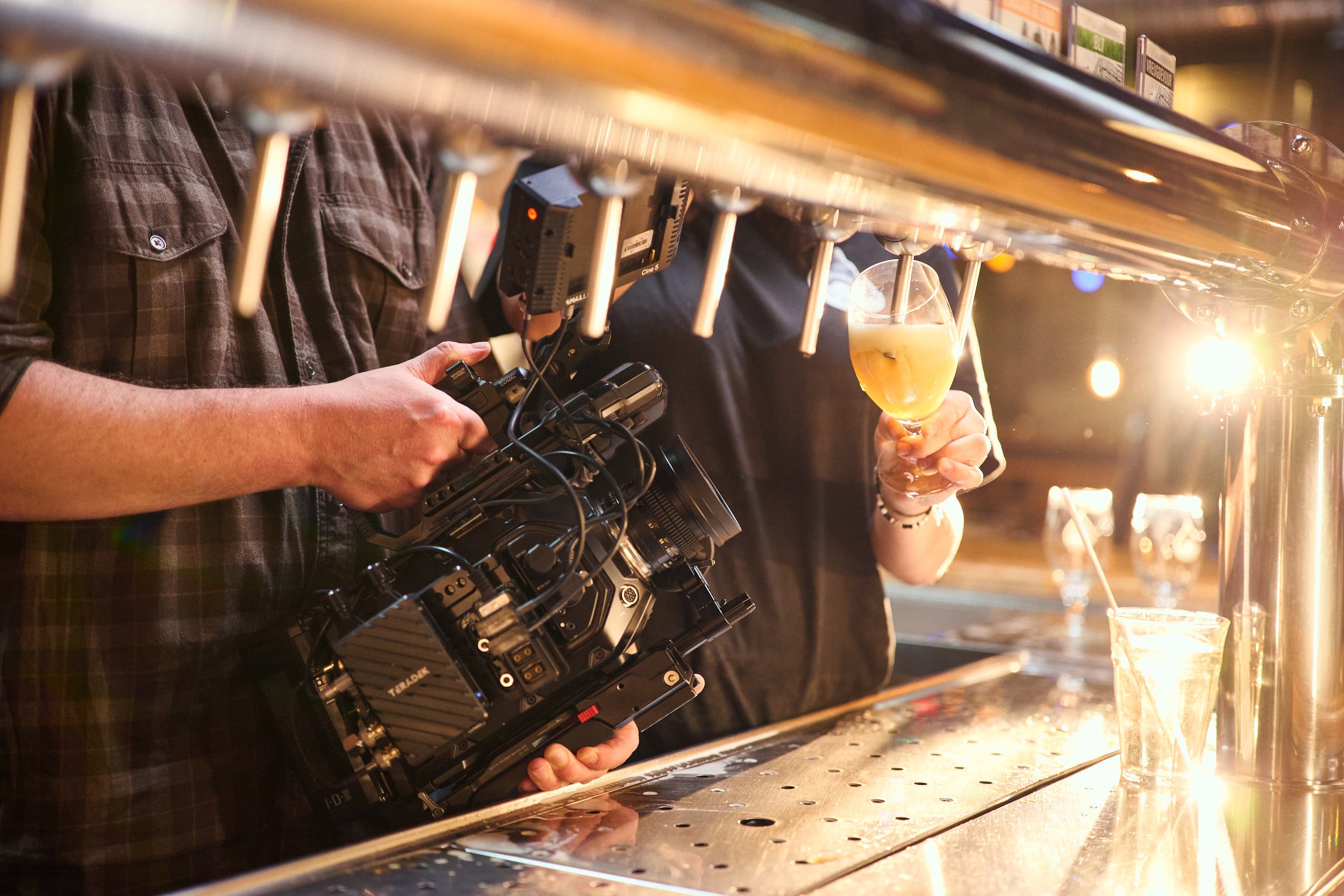 A person holding a professional video camera filming a bartender pouring a drink at a bar with glasses of beer and cocktail glasses on the bar counter.