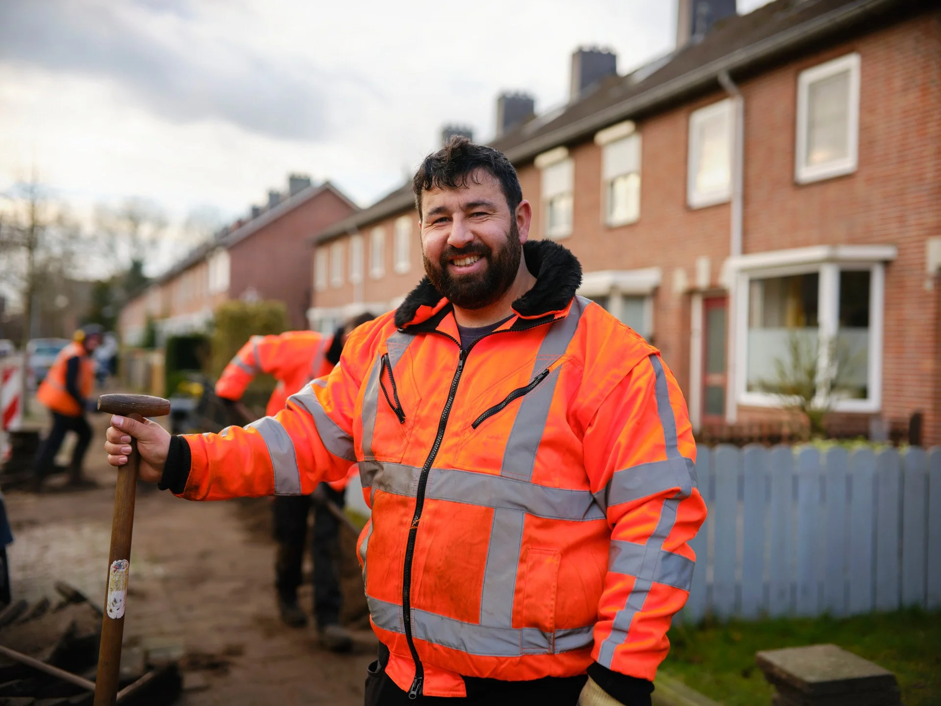 Man in orange safety jacket smiling at construction site with workers and houses in background.