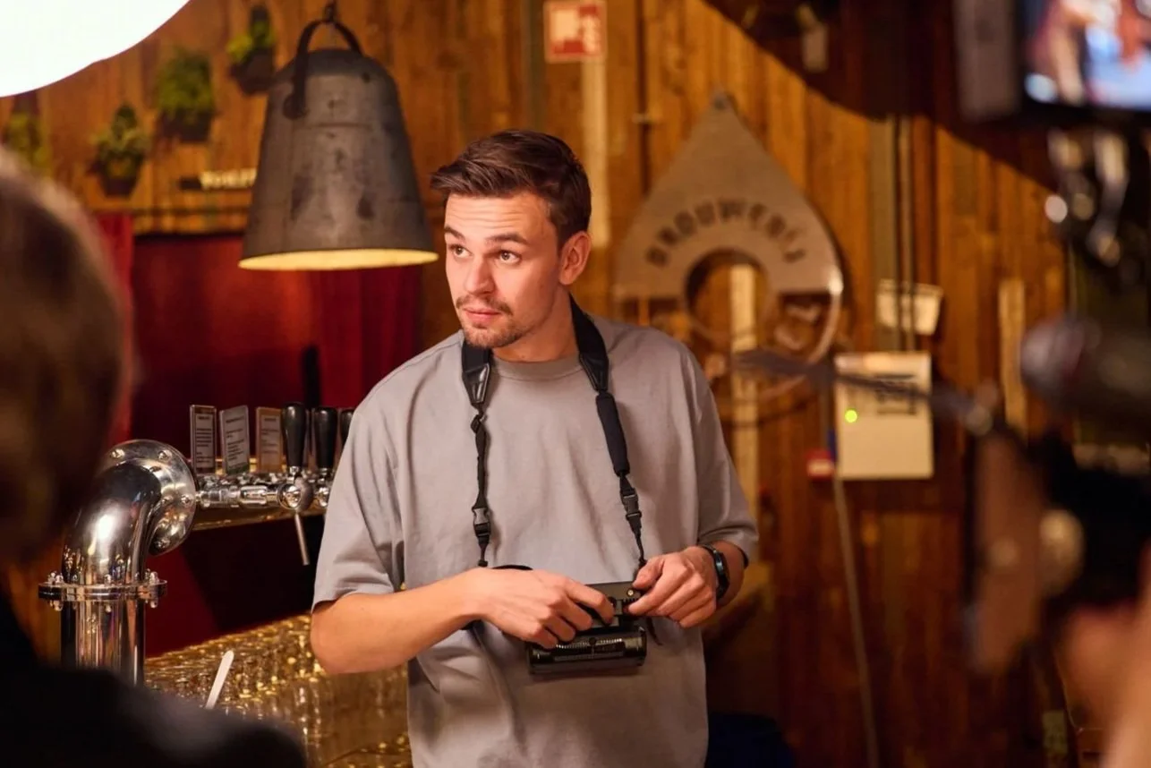 A man with short brown hair and a beard, wearing a gray t-shirt, holding a vintage camera with a strap around his neck, standing inside a rustic bar or brewery with wood-paneled walls and beer taps in the background.