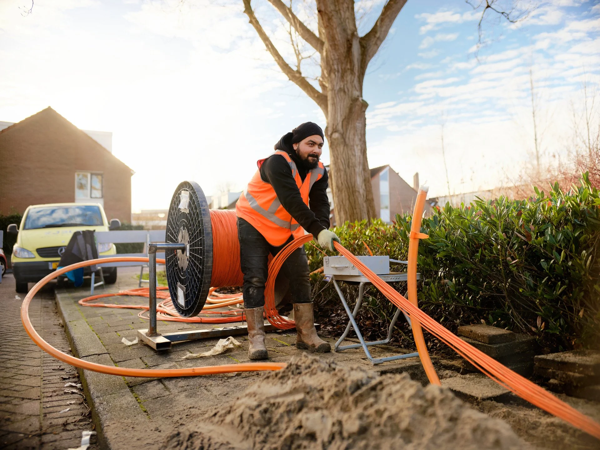 A worker in an orange safety vest and gloves installing underground cables in a trench next to a hedge, with a tree and residential buildings in the background.