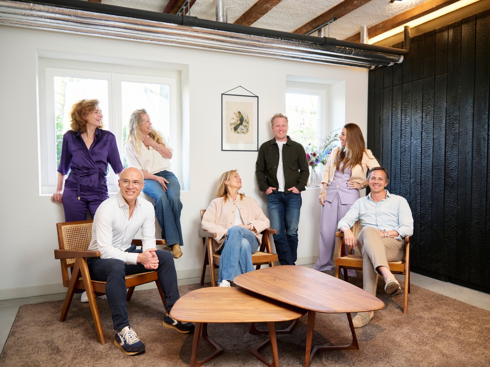 Group of seven people, four women and three men, gathered in a modern room with white walls and black textured wall panel. Two are sitting on wooden chairs, and three women are sitting on the windowsill, with the remaining man and woman standing. The