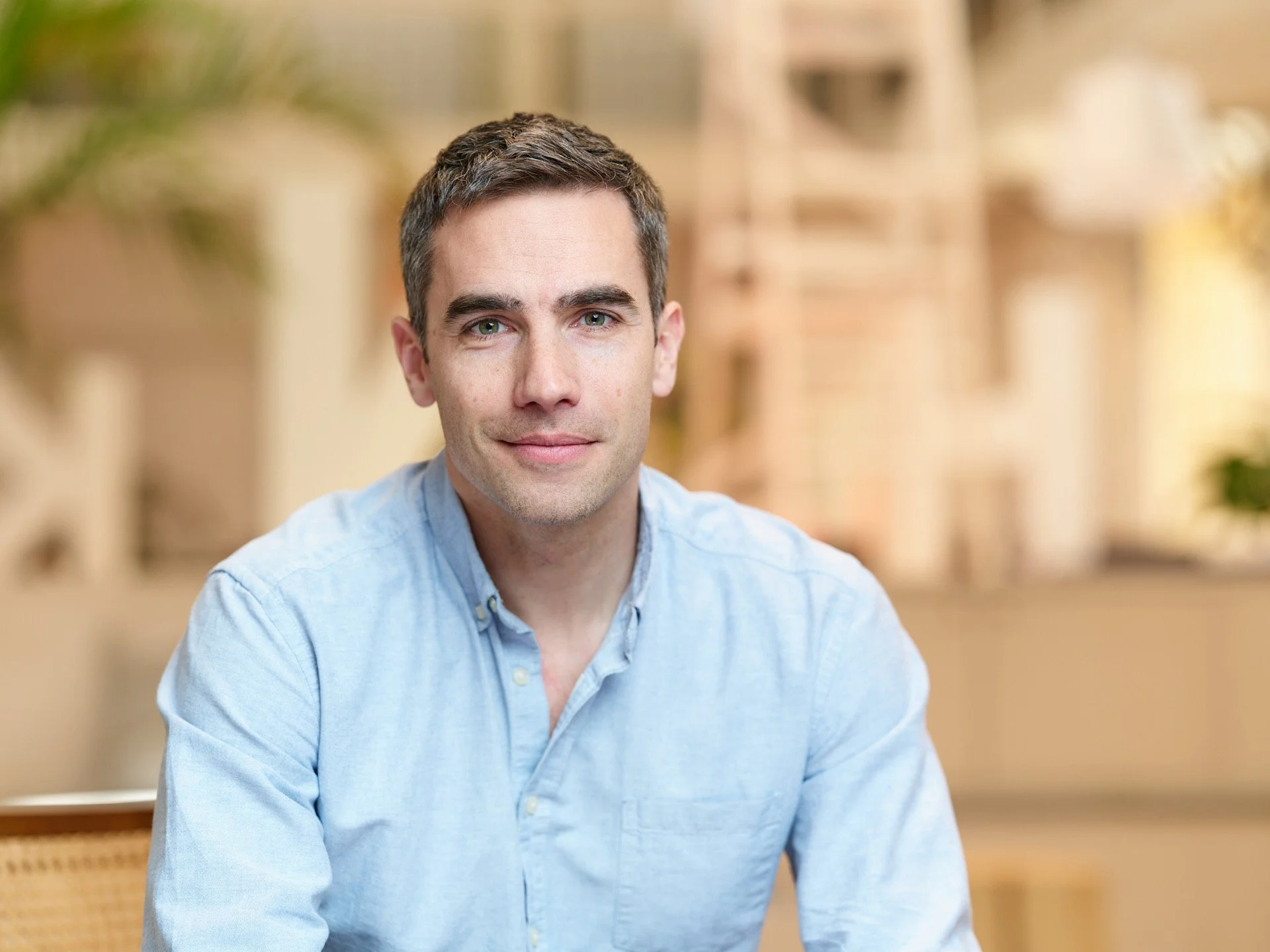 A man with short brown hair and blue eyes, wearing a light blue button-up shirt, sitting indoors with a blurred bookshelf and plants in the background.