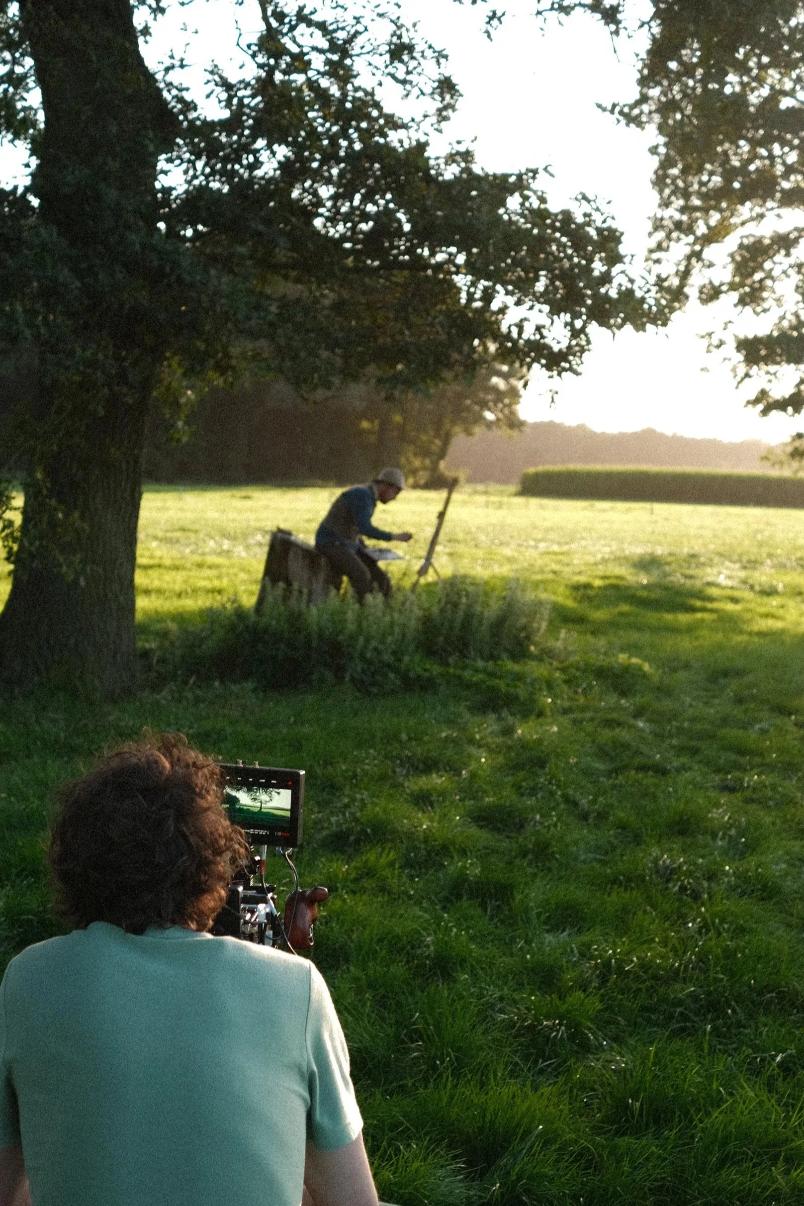 A person with curly hair operating a camera on a tripod, filming a man sitting on a tree stump in a grassy field, shaded by a large tree, during sunset.