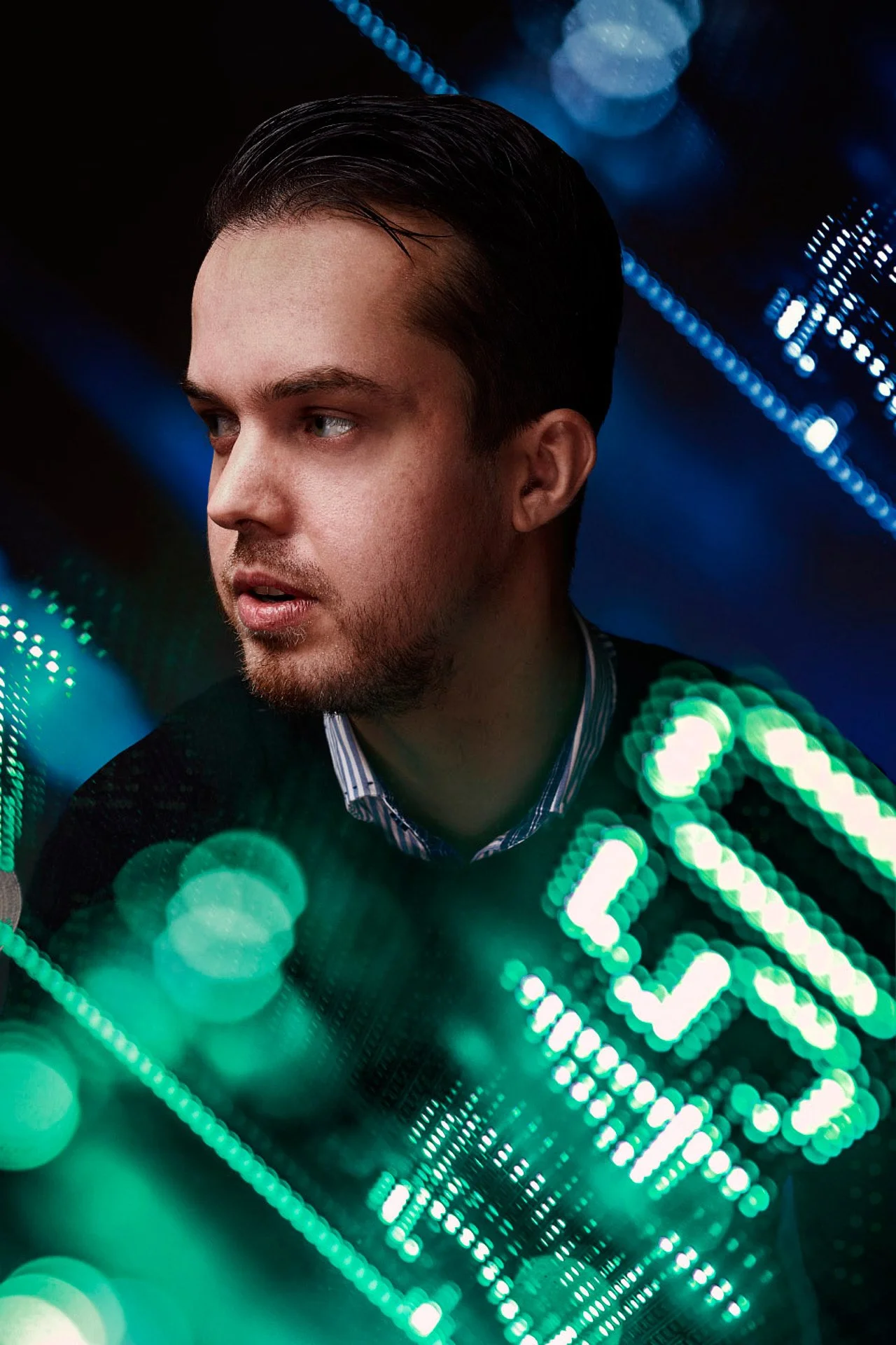 A man with dark hair and a beard looking to the side in front of a background of bright blue and green bokeh lights.