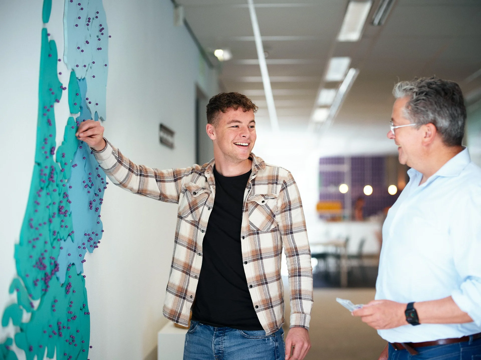 Two men conversing in an office, one pointing at a colorful wall art display.