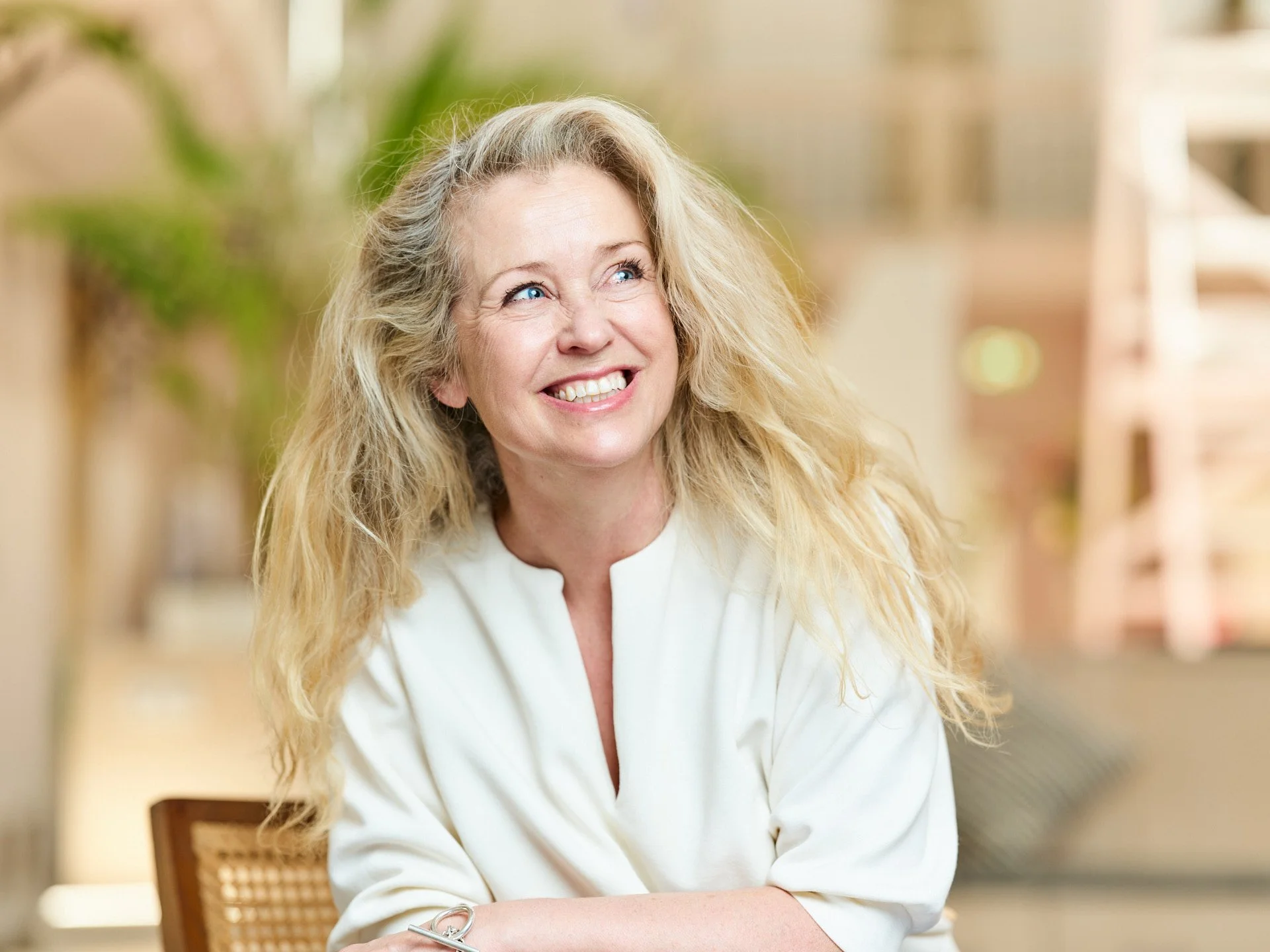A middle-aged woman with blonde, curly hair and blue eyes, smiling and looking up, sitting in a bright indoor or outdoor space with blurred plants and architecture in the background.
