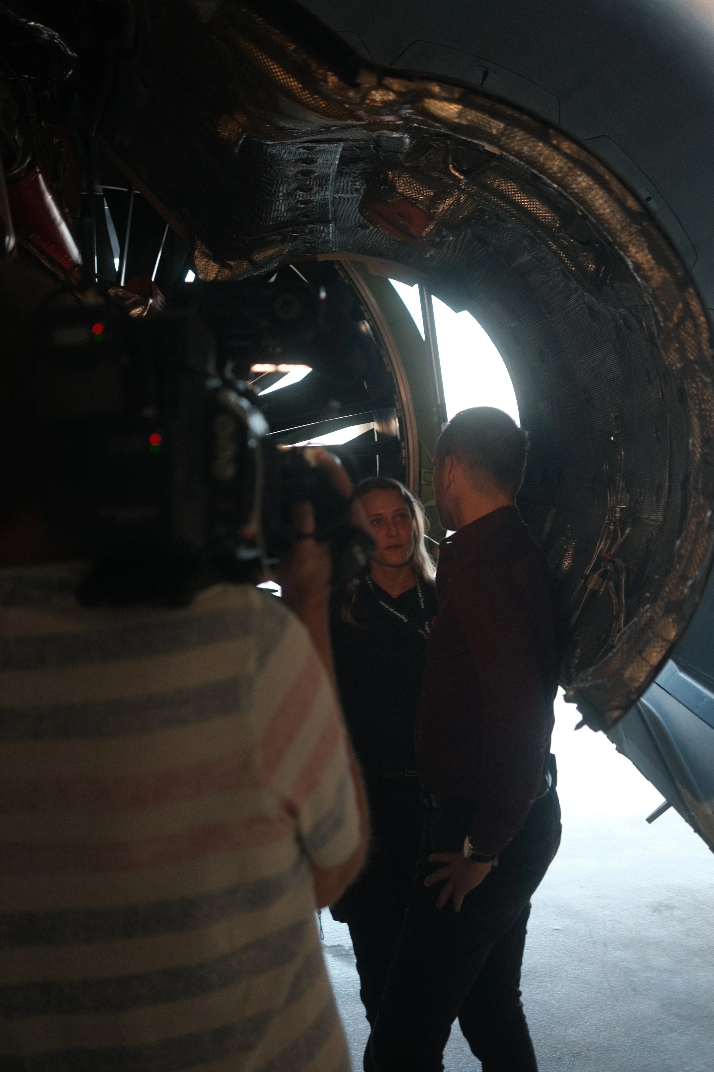 Two people, a man and a woman, talking to each other inside a large metallic structure, with a person in a striped shirt filming or photographing the scene.