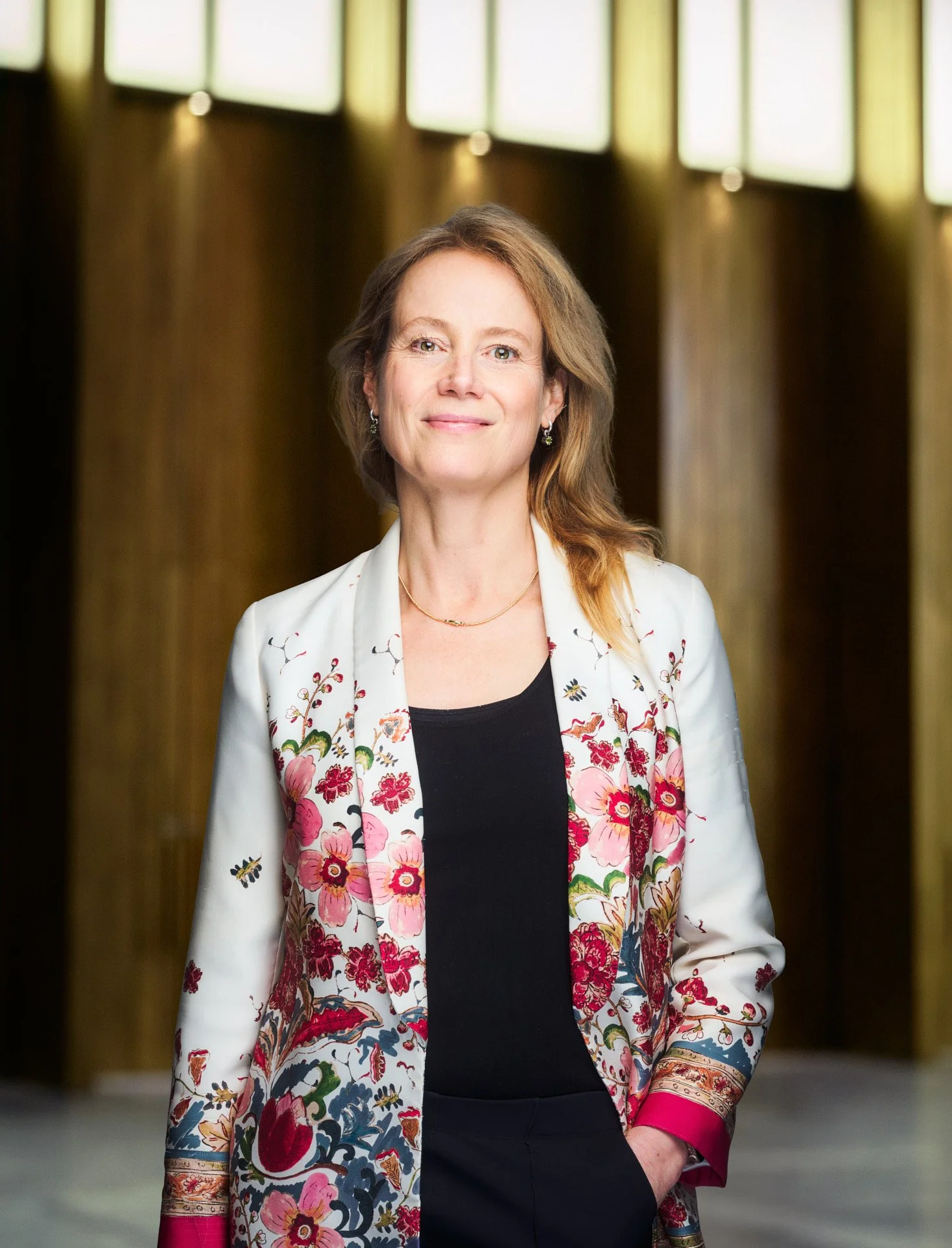 A woman with shoulder-length light brown hair wearing a white floral blazer and black top, standing in a hallway with large windows.