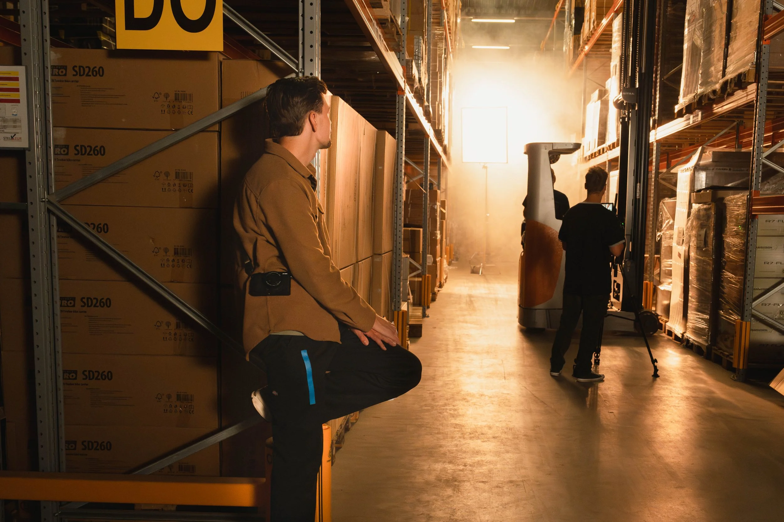 A man leaning against shelves in a warehouse, watching two others work with equipment near bright sunlight.