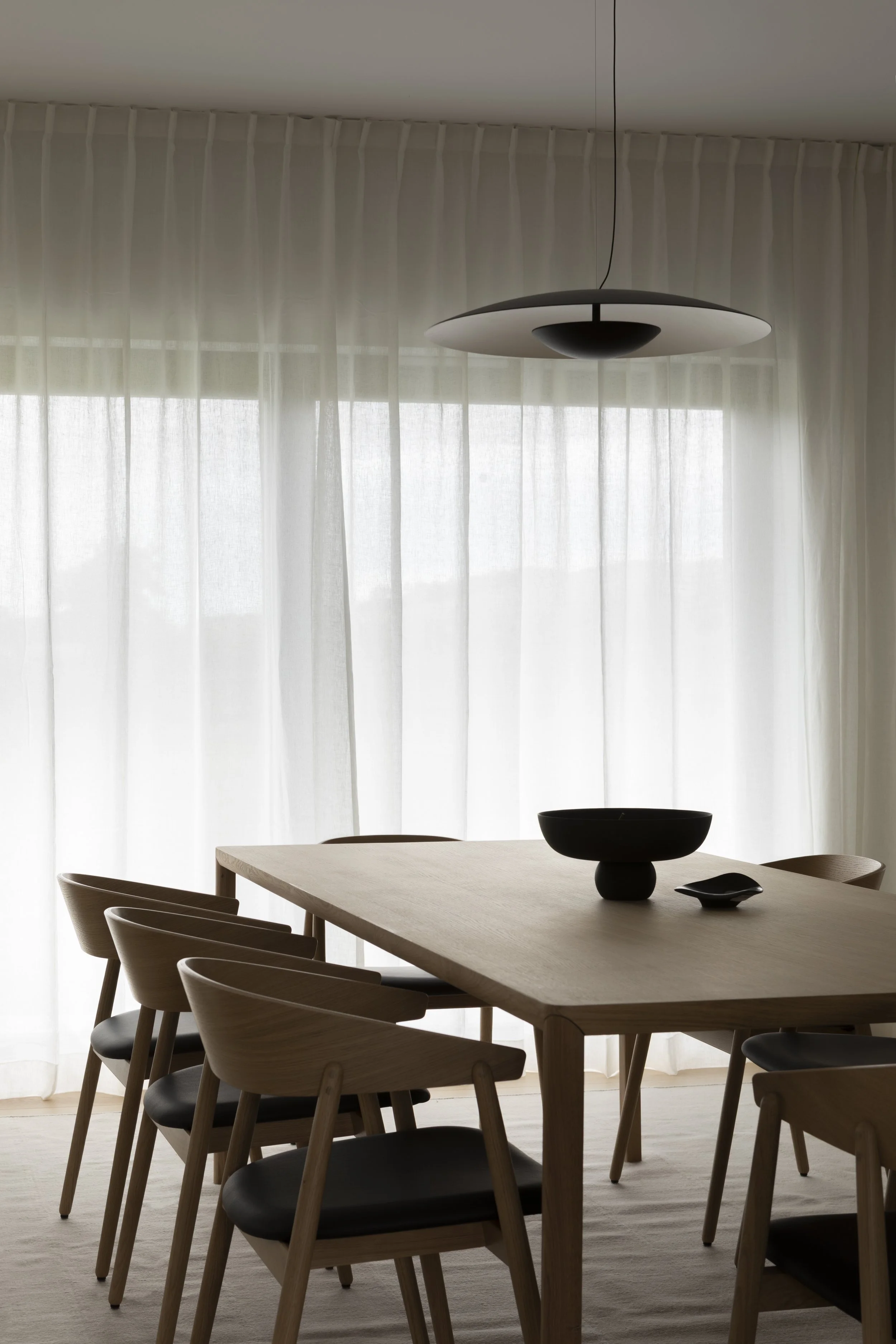 Minimalist dining room with a wooden table, six matching chairs, a black bowl and a small black dish, and a modern hanging ceiling light. Sheer white curtains cover large windows in the background.
