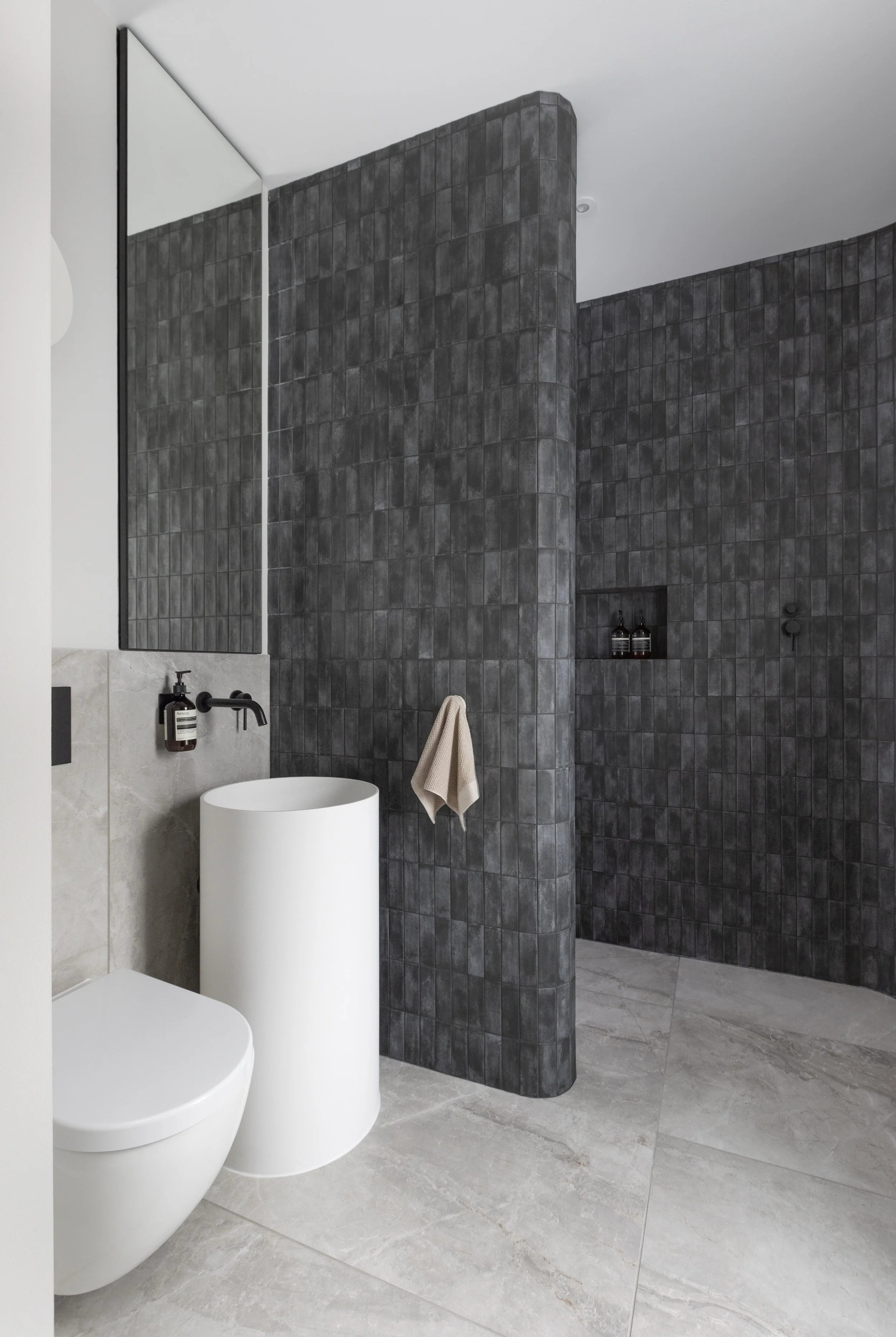 Modern bathroom with a black-tiled shower area and a white freestanding sink, beige towel on the wall, and a large mirror.