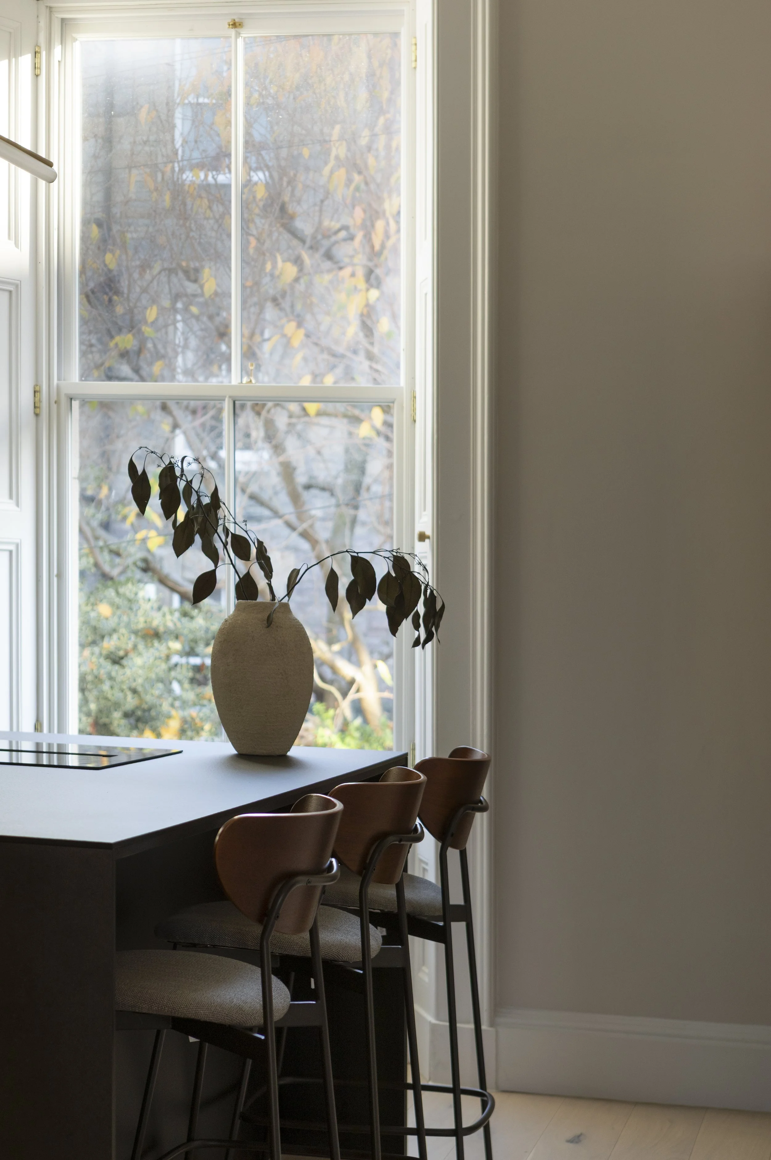 Interior view of a kitchen with a large window, a white countertop with a decorative vase and leafy branch, and three wooden bar stools.