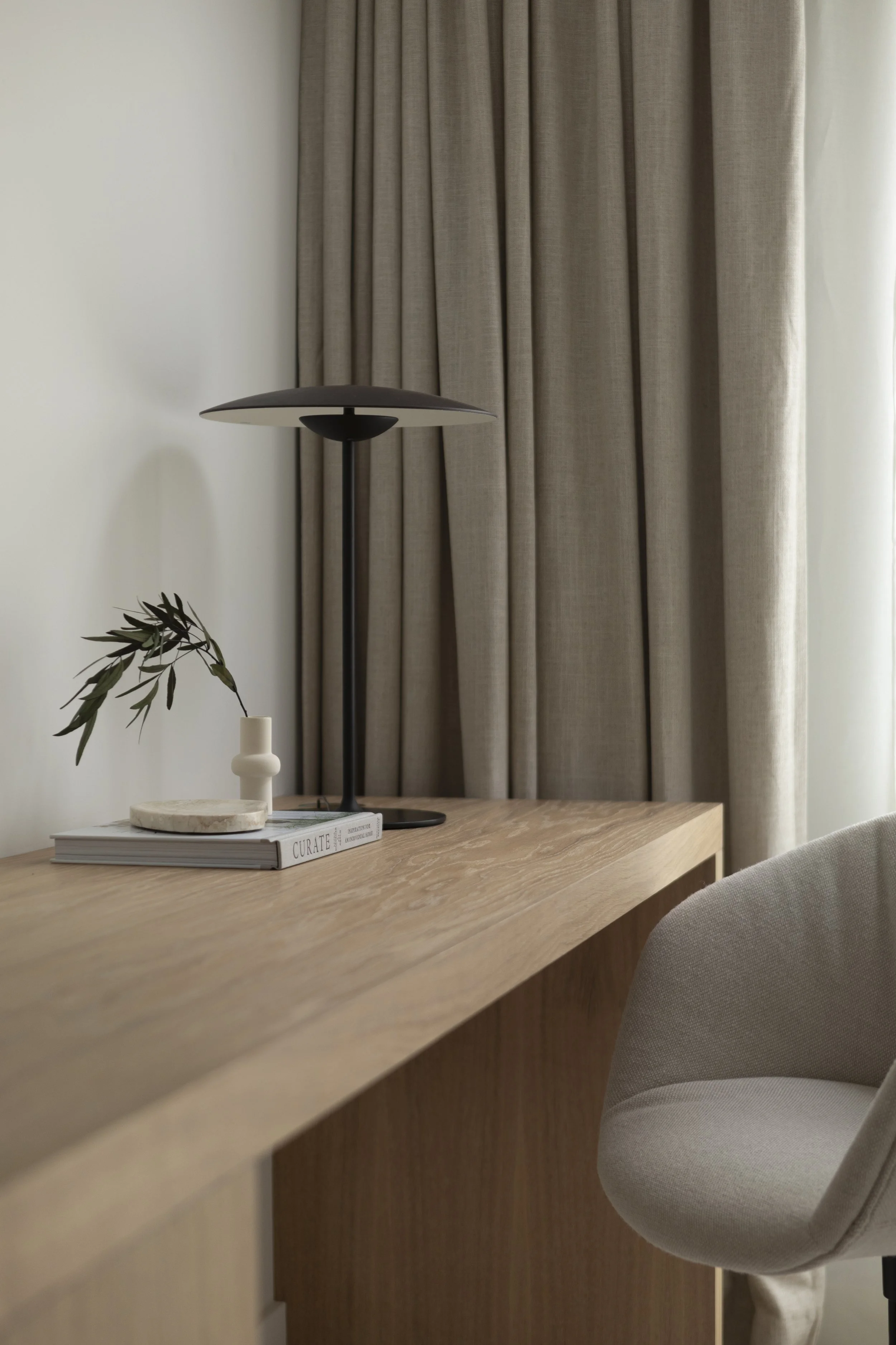 A wooden desk with a black modern table lamp, a small white vase with an olive branch, a stack of books, and a beige upholstered chair in front of beige curtains.