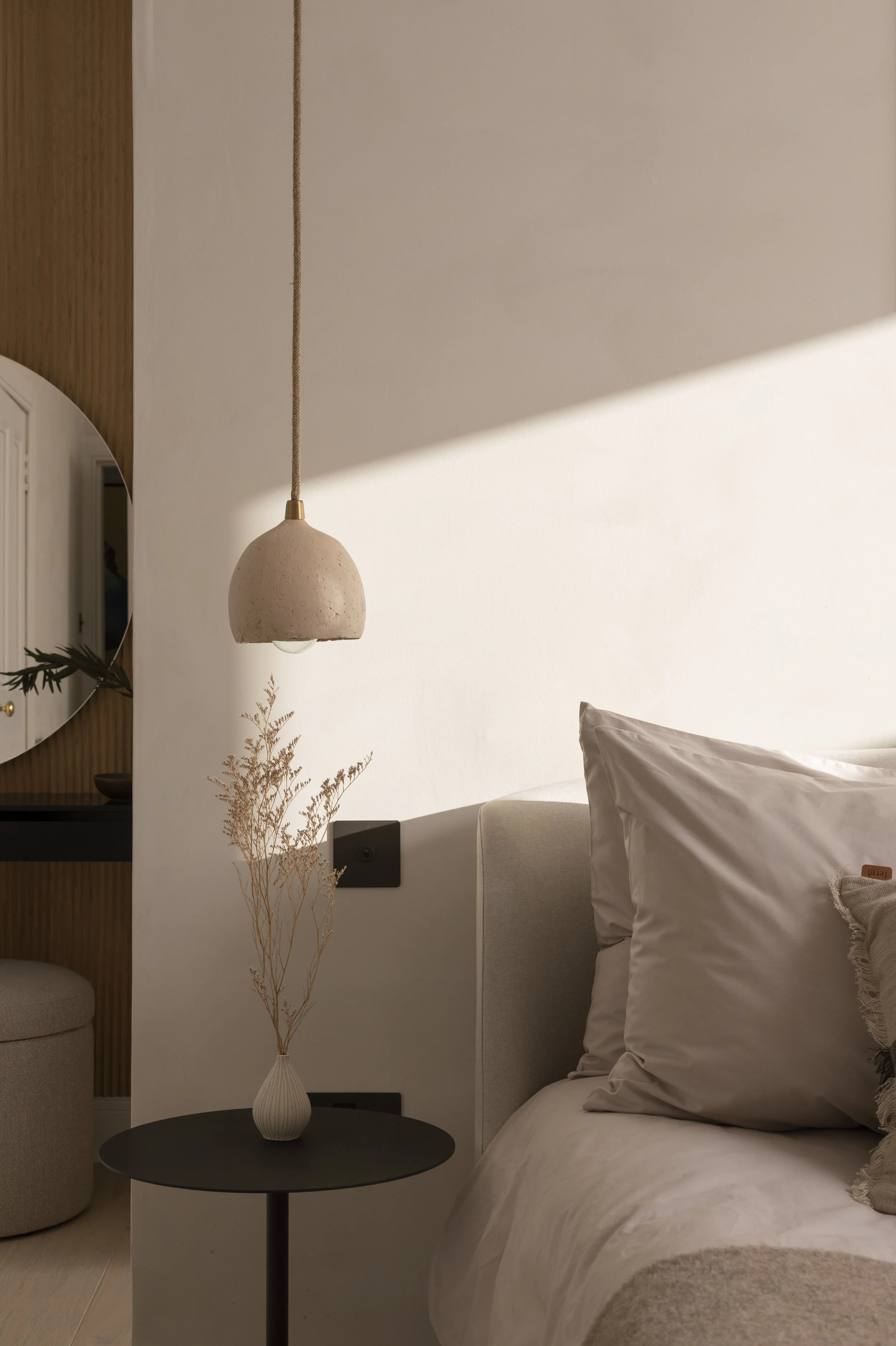 A bedroom with a white wall, beige headboard, and light-colored bed linens. A round black side table with a white vase holding dried branches, a hanging pendant light, and a mirror reflecting part of the room are visible.