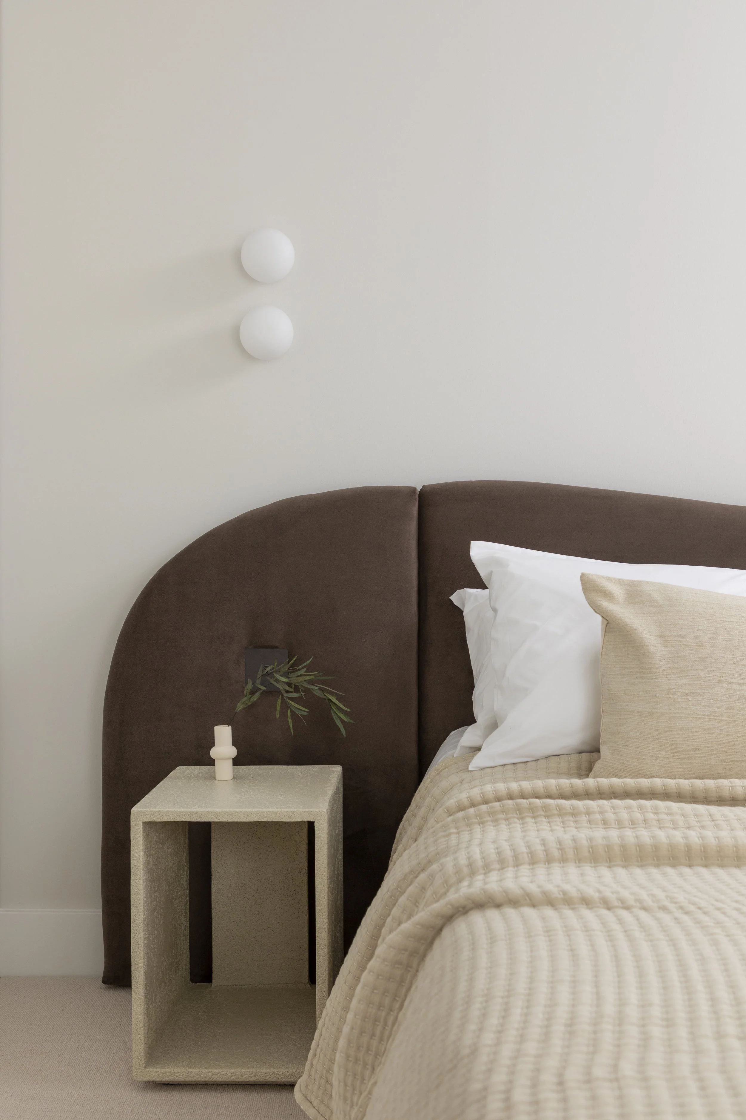 A bedroom with a beige bedspread, white and beige pillows, a dark brown upholstered headboard, a minimalist beige nightstand holding a small plant, and two round white wall sconces on a plain white wall.