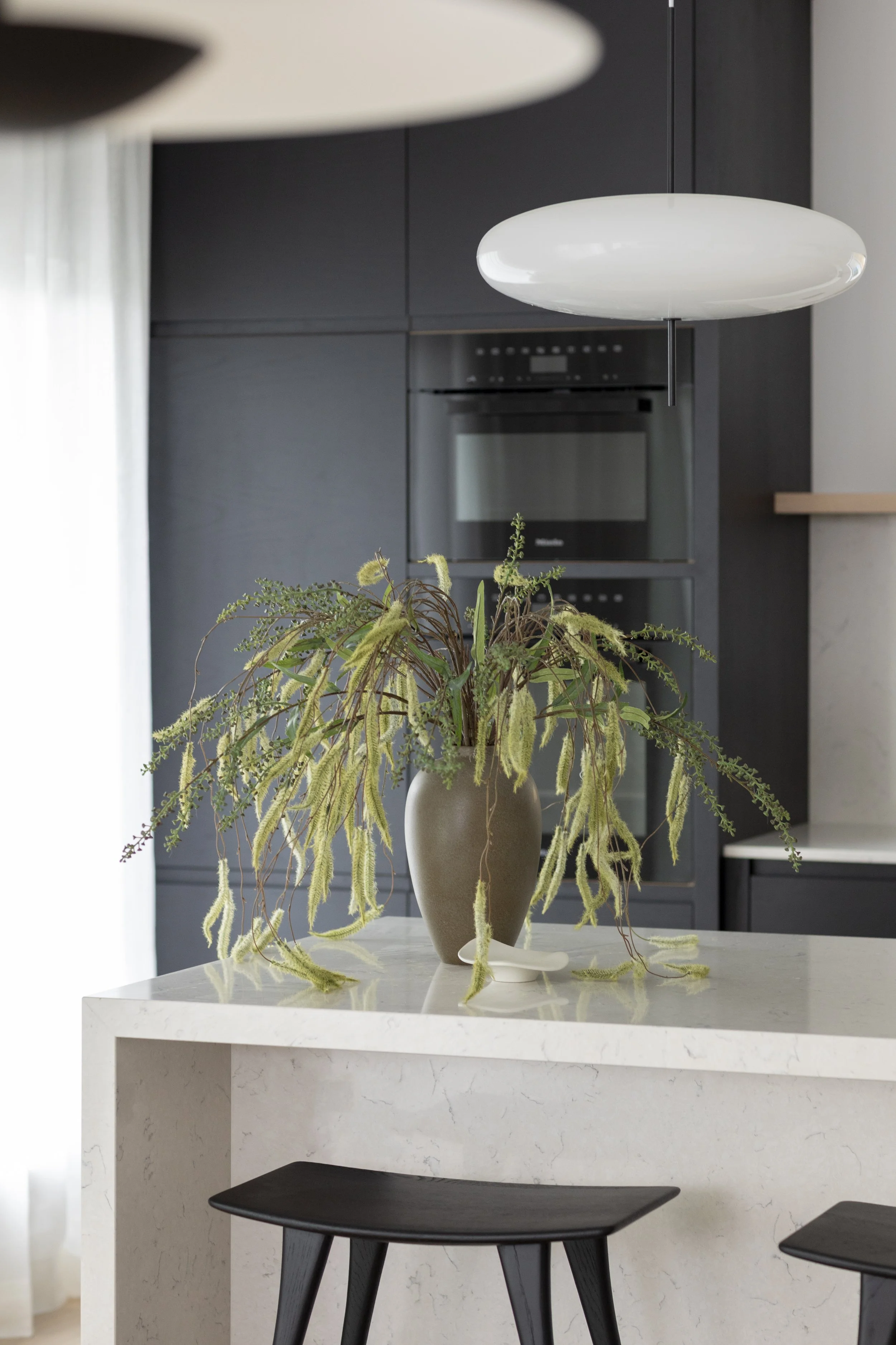 Modern kitchen with a white marble countertop, black chairs, a decorative plant in a beige vase, and black cabinetry with a built-in oven.