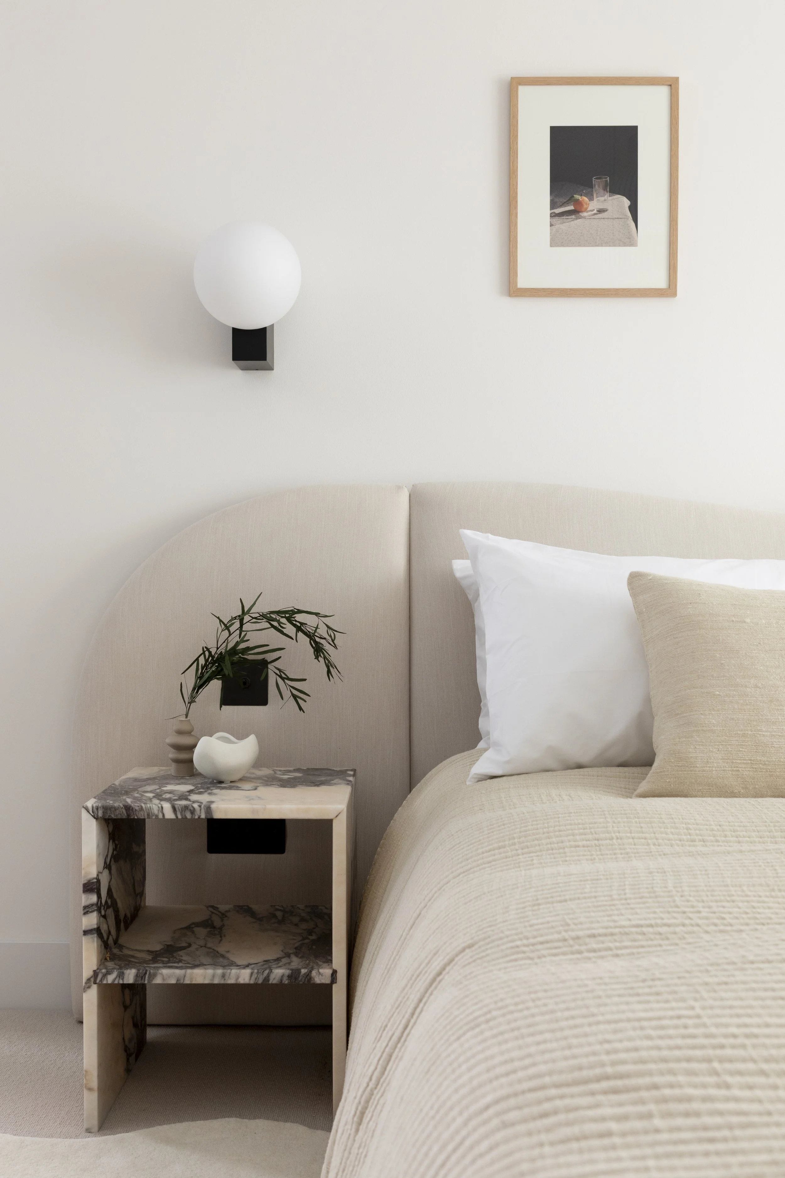 A bedroom with a beige upholstered headboard, beige bedding, white and beige pillows, a marble side table with a small plant, a white decorative object, a wall-mounted black and white artwork, and a wall-mounted white globe light.