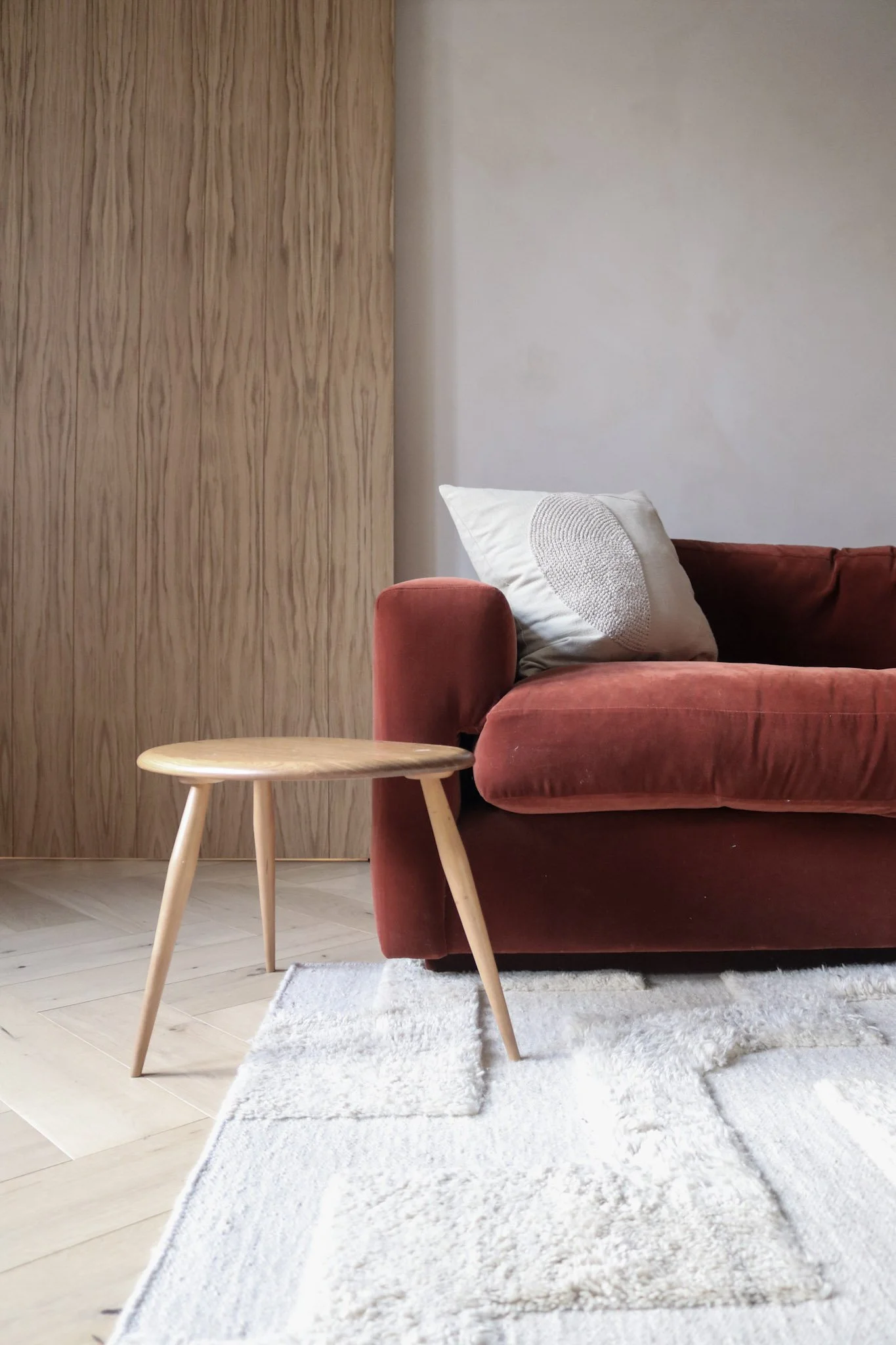 Living room with a red velvet couch, a beige pillow, a small wooden side table, light-colored wood flooring, a white textured area rug, and a wooden wall panel.