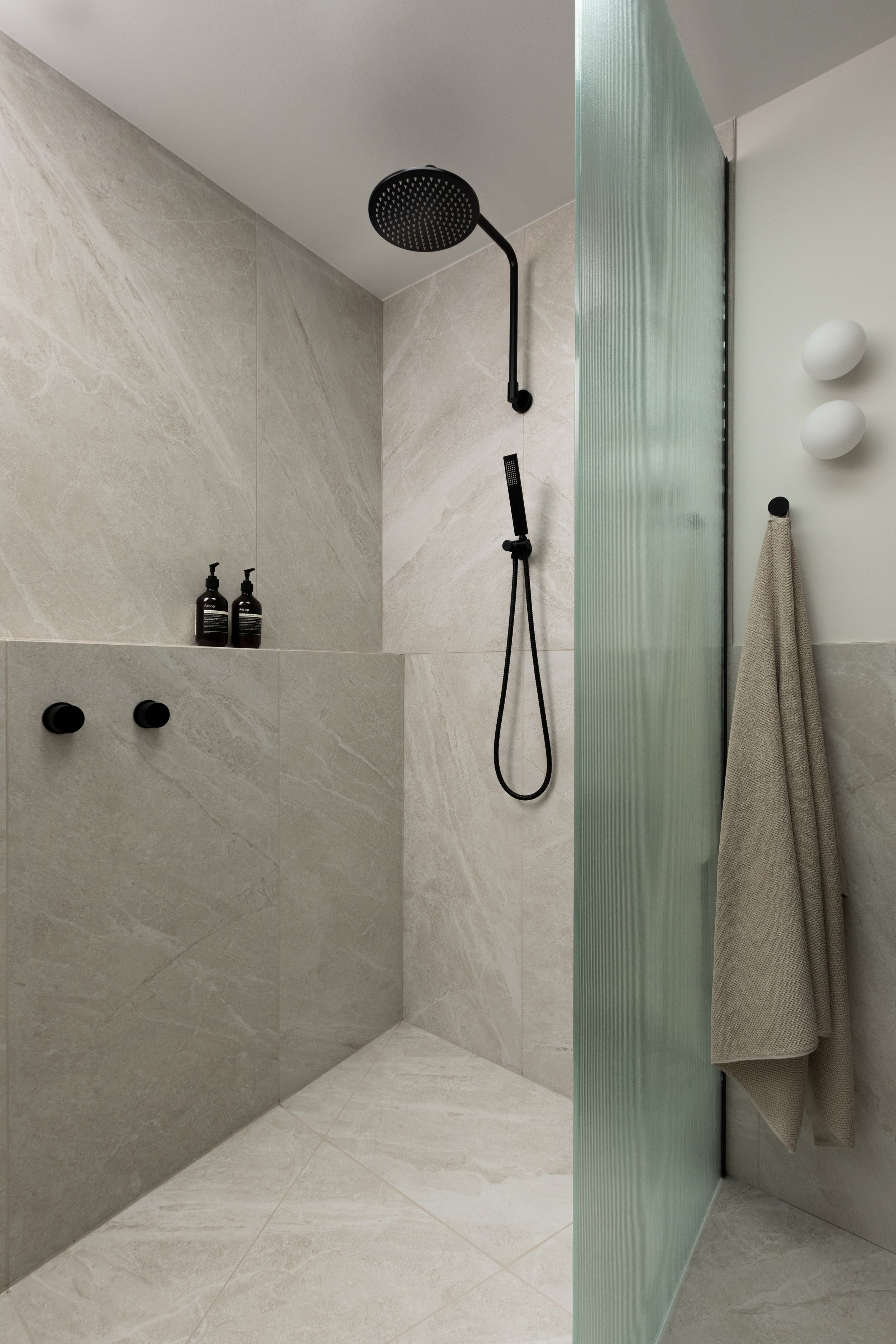 Modern shower with black fixtures, beige tiled walls and floor, a frosted glass partition, a towel hanging on a hook, and two wall-mounted white spherical lights.