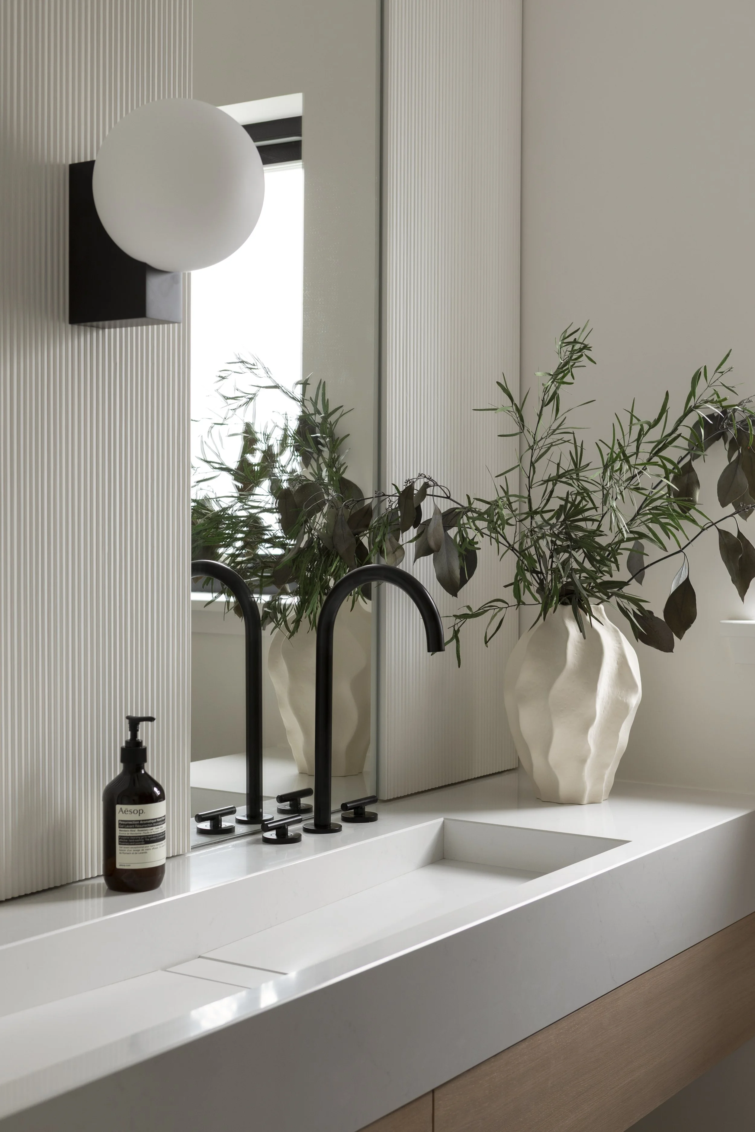 Modern bathroom vanity with a white sink, black faucet, a mirror, and two large vases with leafy green plants near a window.