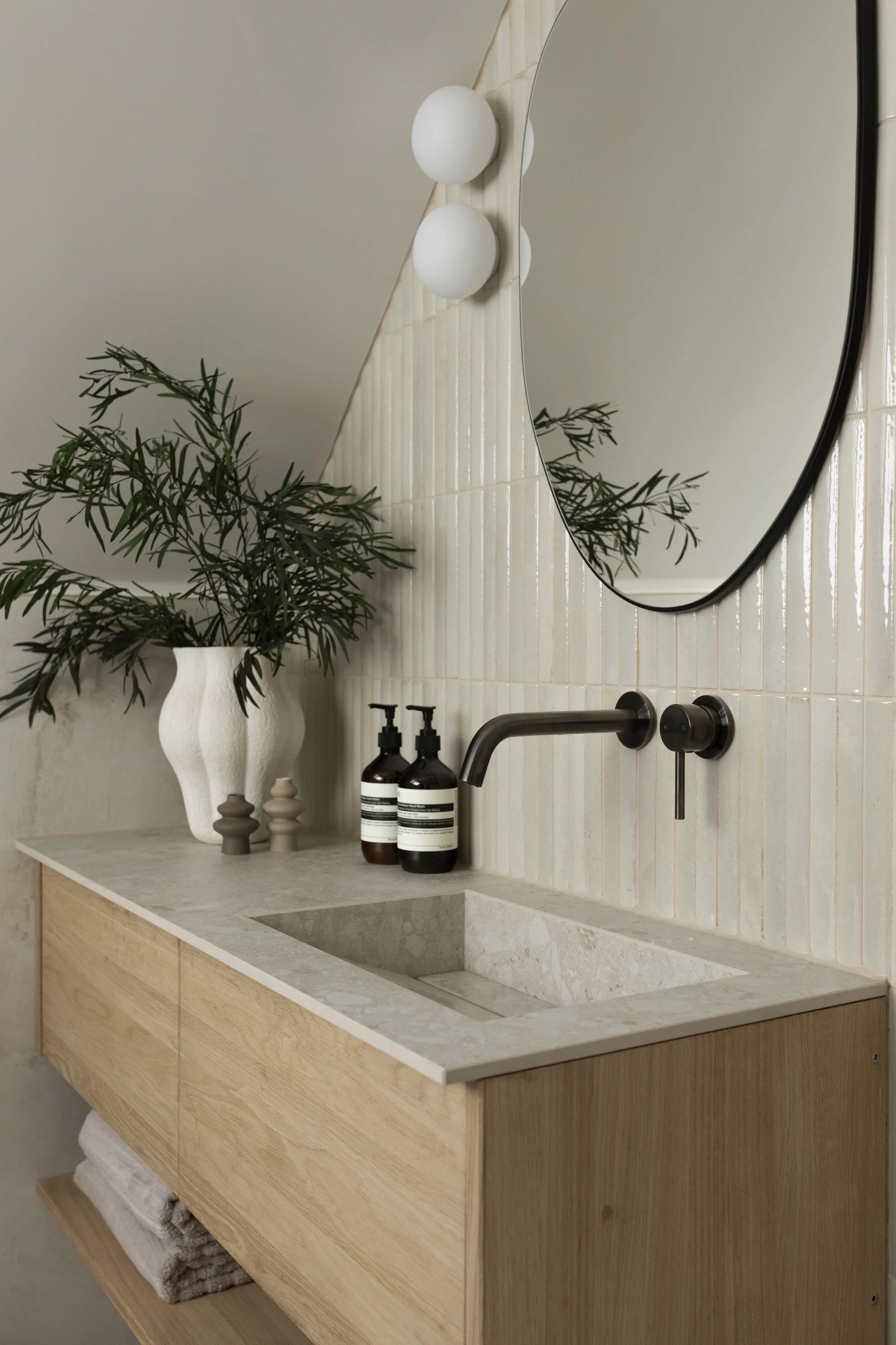 Modern bathroom vanity with a rectangular stone sink, a wooden cabinet, a large round mirror, a potted plant, and two bottles of soap or lotion.