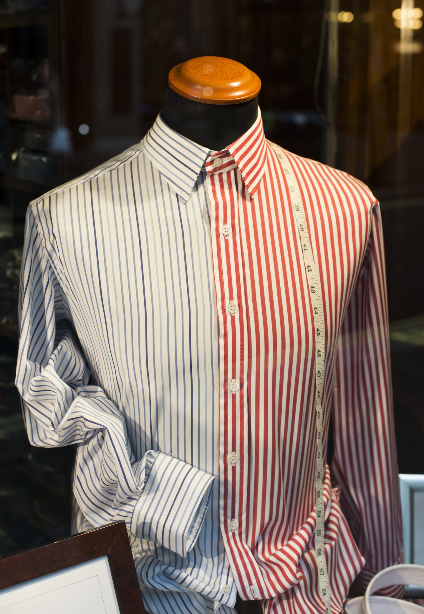 A mannequin wearing a button-up shirt with red and white vertical stripes on the right side and blue and white vertical stripes on the left side, with a measuring tape draped over the shoulder.