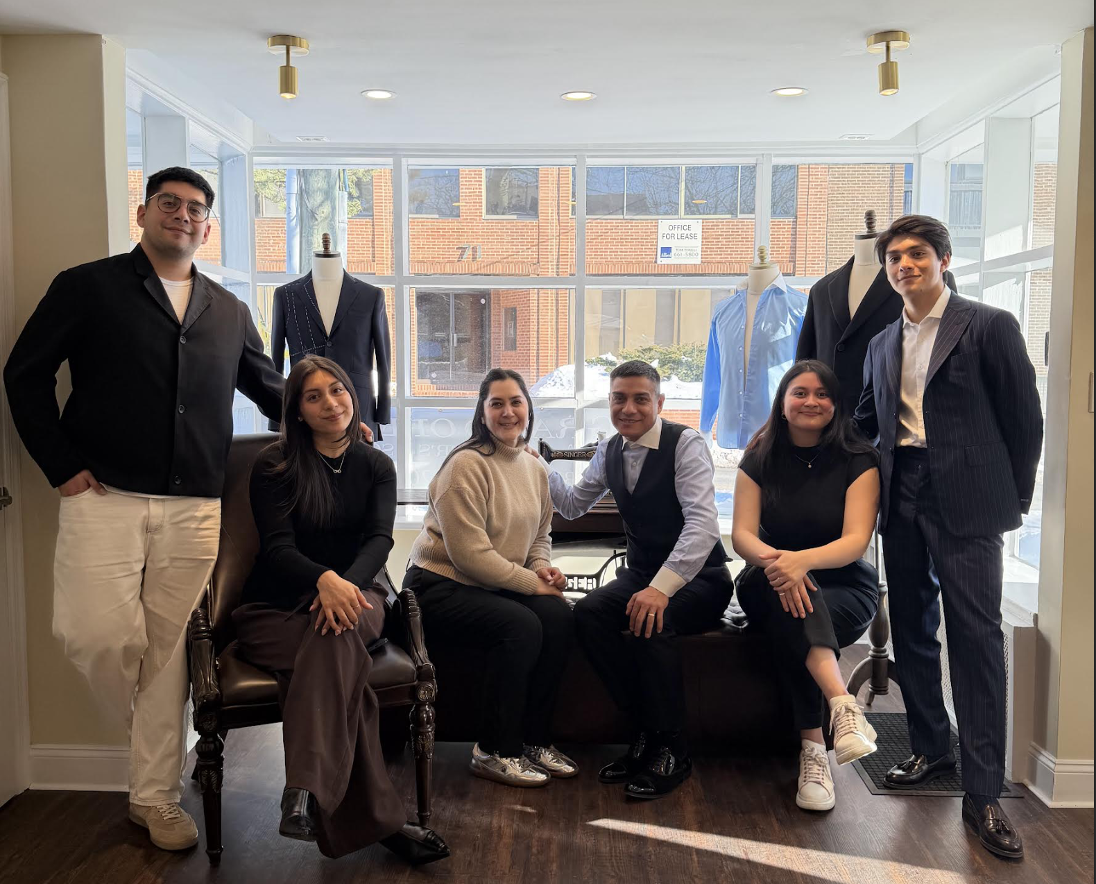 Group of seven young adults in a modern retail store or showroom with mannequins dressed in formal and casual clothing, posing for a photo by large windows with an office building visible outside.