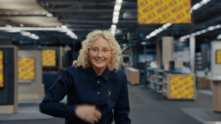 A smiling woman with curly blonde hair and glasses standing in an electronics store with shelves and display screens in the background.