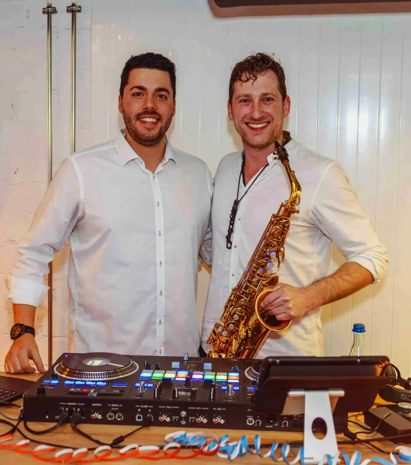 Zwei Männer, die neben DJ-Equipment stehen, Frederik Heckel mit Saxophon, beide lächeln in die Kamera, weiße Hemden, hinter ihnen eine weiße Wand.