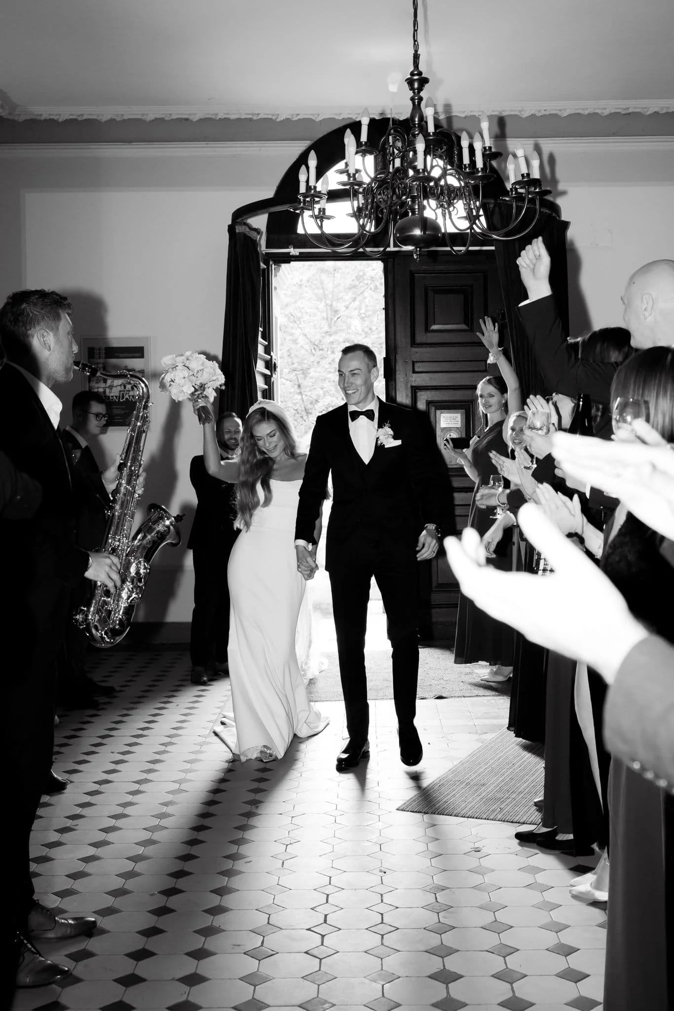 Schwarz-weiß-Foto von einem frisch verheirateten Paar, das die Tür eines Raumes betritt, umgeben von jubelnden Gästen, einigen mit Champagner. Frederik Heckel mit Saxophon steht links.